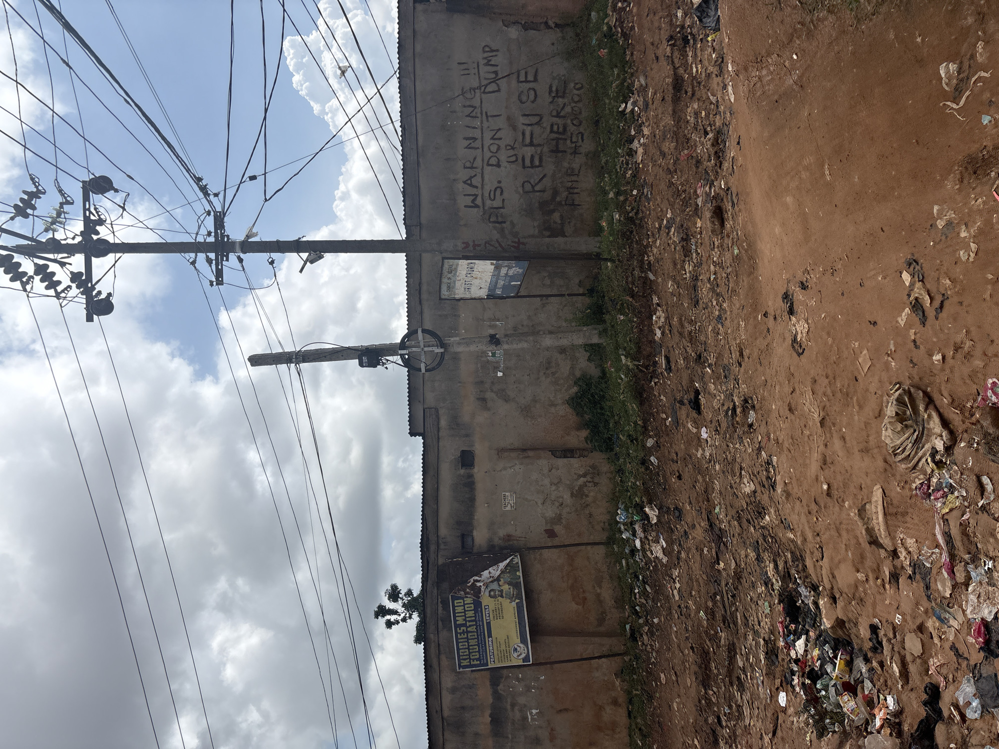 The image depicts an outdoor scene in Nkpor, Nigeria, characterized by a dusty, unpaved ground littered with various types of refuse, including plastic bags, bottles, and other debris. A weathered concrete wall stands prominently, serving as a backdrop. On this wall, graffiti in black paint reads "WARNING!!! PLS. DON'T DUMP UR REFUSE HERE FINE #50000." Below this warning, a torn poster advertises "KIDDIES MIND FOUNDATION," listing different educational levels offered and mentioning "TEACHERS."
The environment appears somewhat neglected, with overgrown green vegetation visible on the left side of the wall. Overhead, a complex network of power lines crisscrosses the sky, supported by utility poles. The sky is partly cloudy, suggesting a daytime setting, possibly in the morning or afternoon. The overall impression is one of a utilitarian and somewhat impoverished urban setting, where regulations against dumping are visibly enforced through signage, and community services like a foundation are advertised. The scene is devoid of people.