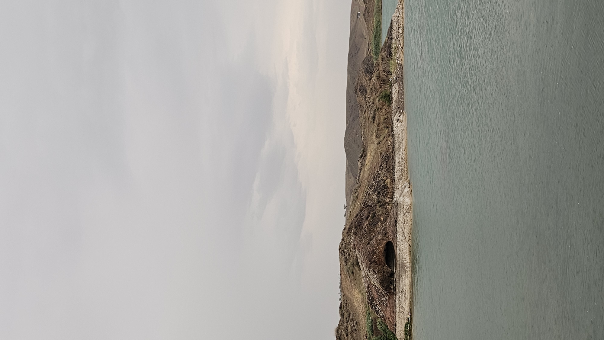 This is a vertical, eye-level, medium shot of a rugged, arid landscape on the shore of a large body of water. The sky is overcast with light gray clouds. The foreground is dominated by a body of blue-green water, appearing somewhat choppy with small ripples and faint white specks that may indicate light rain. The middle ground features a rocky, brown-hued hillside with sparse vegetation. A notable feature is a dark, cave-like opening visible in the side of the hill. The terrain rises into rolling hills in the background, also covered in dry grass and soil. A few scattered structures, possibly houses or buildings, are faintly visible on the distant hills. The overall impression is one of a natural, somewhat desolate environment. The image is located in the city of Qonaev, Kazakhstan.