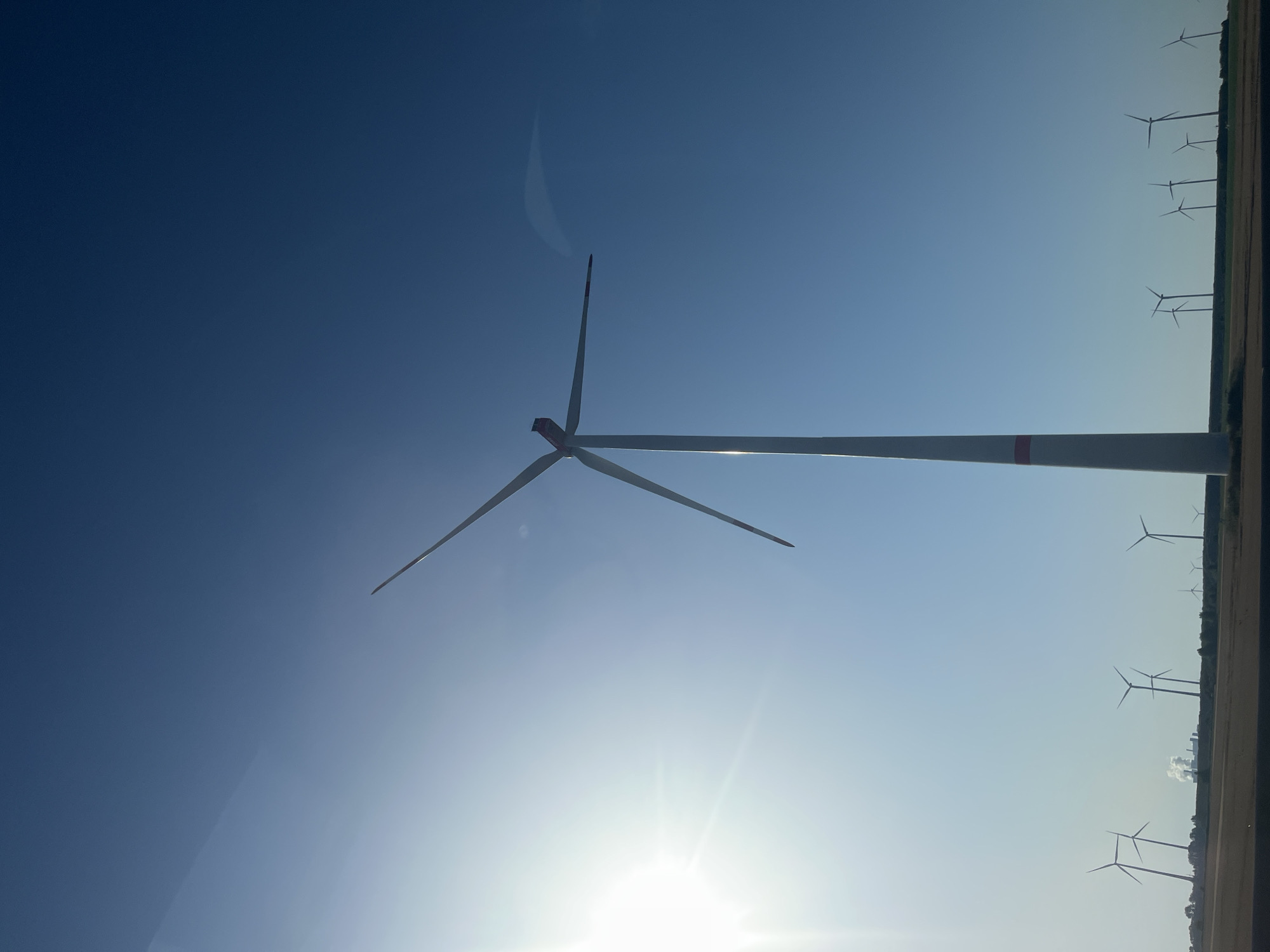 The image shows a wind farm in the city of Jüchen, Germany, under a clear blue sky. A large, prominent wind turbine stands in the foreground, its blades angled upwards. In the background, a row of smaller wind turbines are silhouetted against the horizon, along with distant industrial structures emitting smoke. The sun is shining brightly on the left side of the frame, casting a lens flare. The ground appears to be a field of dry vegetation. There are no people visible in the image.
