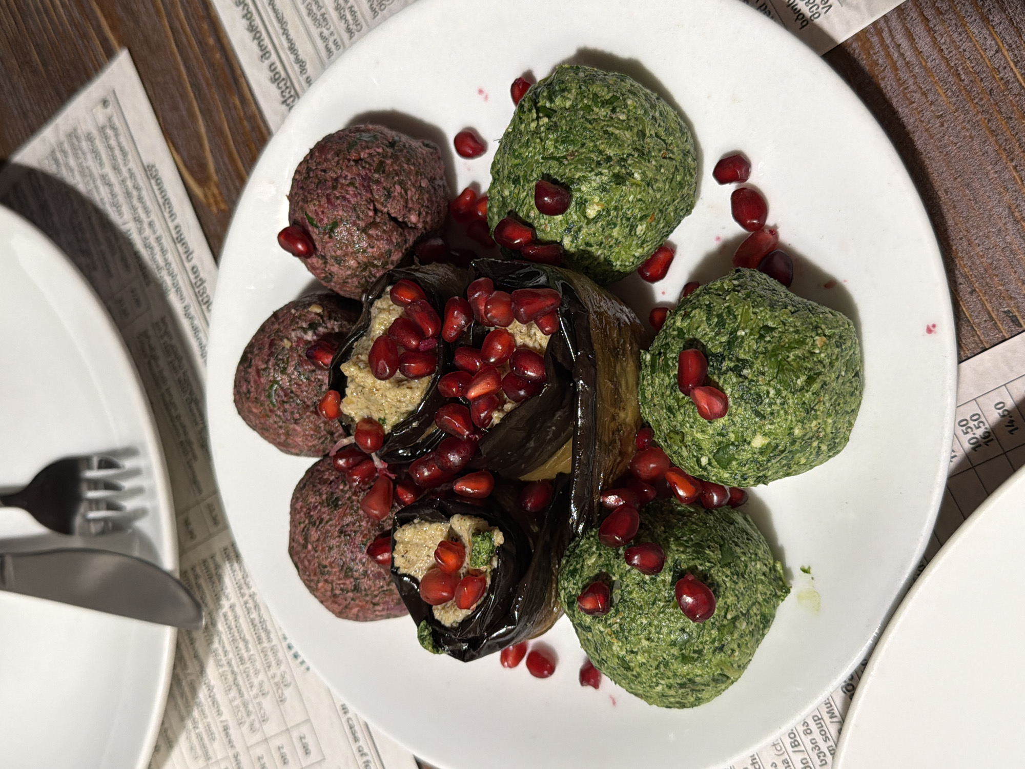 The image shows a plate of Georgian food, likely found in a restaurant in Tbilisi, Georgia. On a white plate are several items: three green spheres, likely spinach pkhali; three darker red spheres, possibly beet pkhali; and eggplant rolls filled with a walnut paste, garnished with pomegranate seeds. The plate is surrounded by a newspaper, parts of which have Georgian text and prices listed. A fork and knife are visible next to a white plate on the left. The table appears to be made of dark wood.