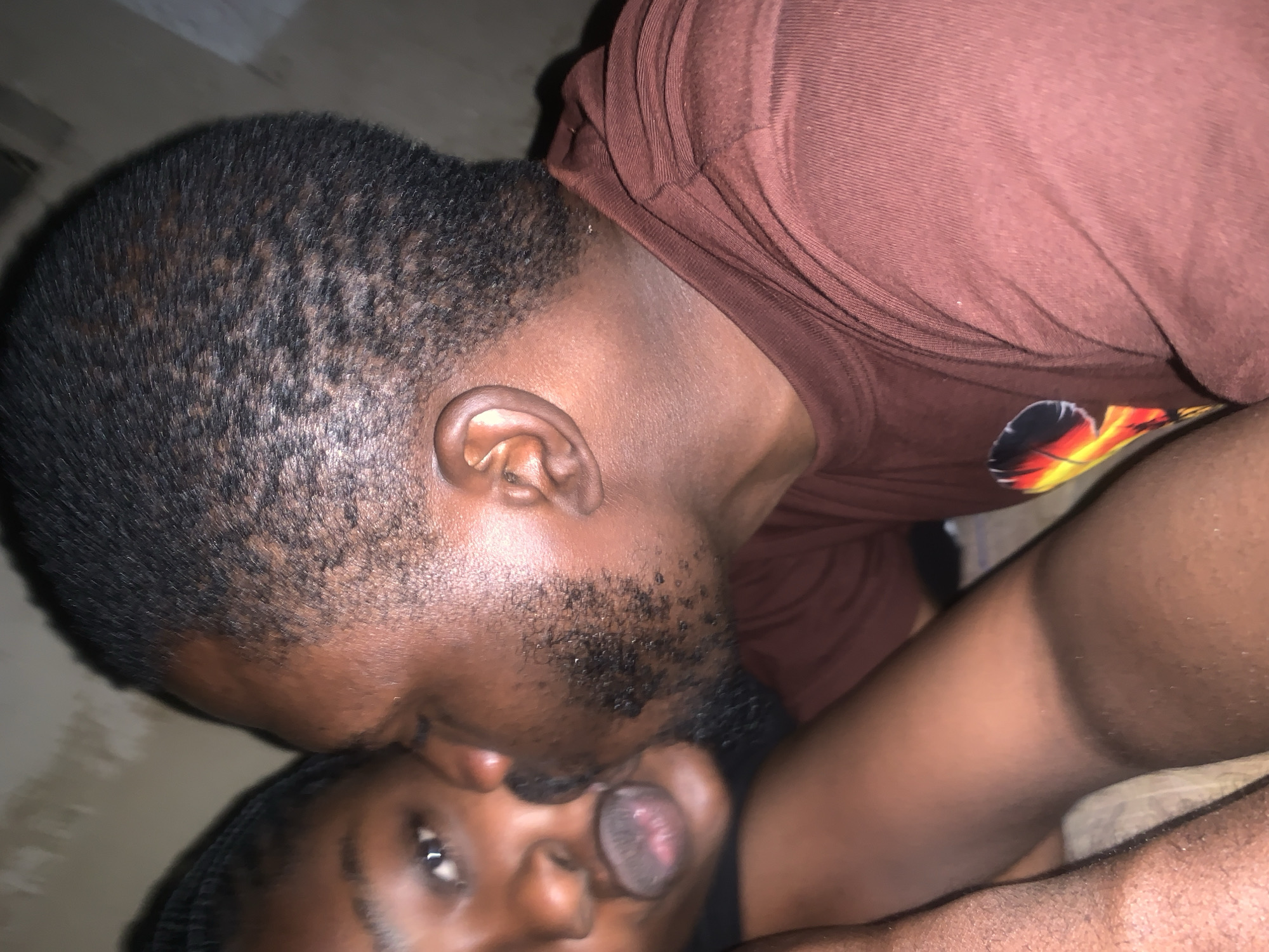 A close-up, slightly blurry image captures two individuals in an intimate moment. A man with short, dark hair and stubble is leaning in to kiss a woman on the cheek. The woman's eyes are visible, looking towards the camera, and her lips are slightly parted. Both individuals have dark skin tones. They appear to be wearing casual clothing, with the man visible in a maroon t-shirt. The background is dimly lit and appears to be an indoor setting, possibly a bedroom, with a textured wall. The overall impression is of a private, affectionate exchange. The image is undated and its location within New Stadium, Nigeria, is not explicitly indicated.