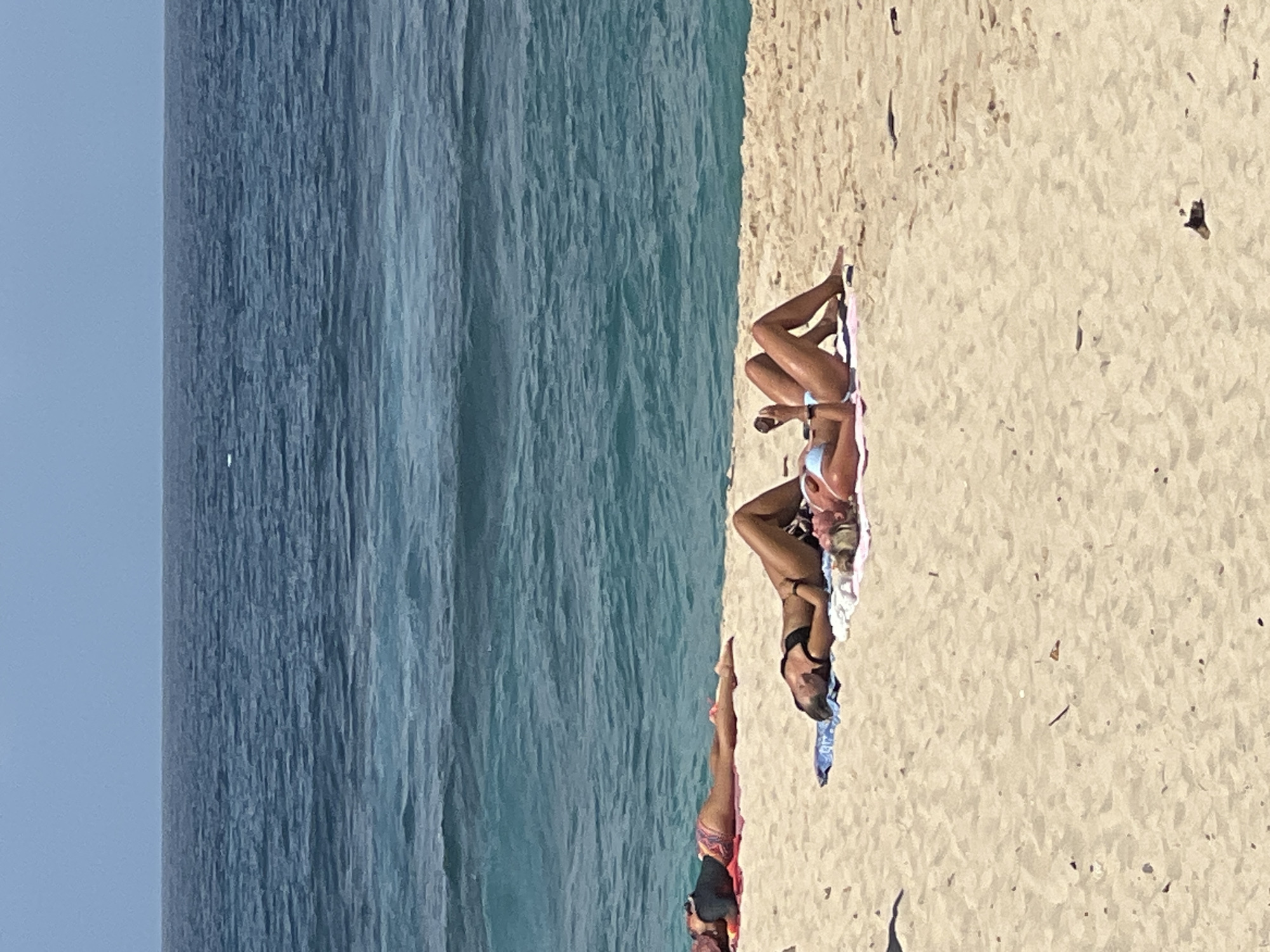 The image depicts a serene beach scene in bright daylight, likely in Kuta Selatan, Indonesia, given the context. The frame is dominated by a clear, light blue sky on the left, transitioning to a vast expanse of turquoise and deep blue ocean in the middle and across the upper left. The ocean appears relatively calm with subtle textures on its surface, indicating gentle movement.
To the right and bottom of the image is a wide stretch of golden-beige sand, characteristic of a tropical beach. Three individuals, all women, are visible relaxing on the sand, suggesting a leisure activity like sunbathing.
In the foreground, a person's legs and lower torso are visible, lying on a colorful patterned towel. They appear to be wearing dark swimwear with some colored details. Further up the beach, near the center-right, a woman with dark hair is lying on her stomach, facing the ocean. She is wearing a black bikini and has sunglasses on her face, resting on a blue towel. Her posture suggests she is relaxing or sunbathing. Slightly behind her, towards the upper right of the beach area, another woman with dark hair is also lying on her stomach, facing the ocean. She is on a light-colored, possibly pink and white striped towel, wearing a light-colored (perhaps white or pale blue) bikini. A small dark object, possibly a phone or a bottle, is visible near her hand.
The overall atmosphere is peaceful and relaxed, typical of a sunny day at a tropical beach. No visible text can be discerned in the image.