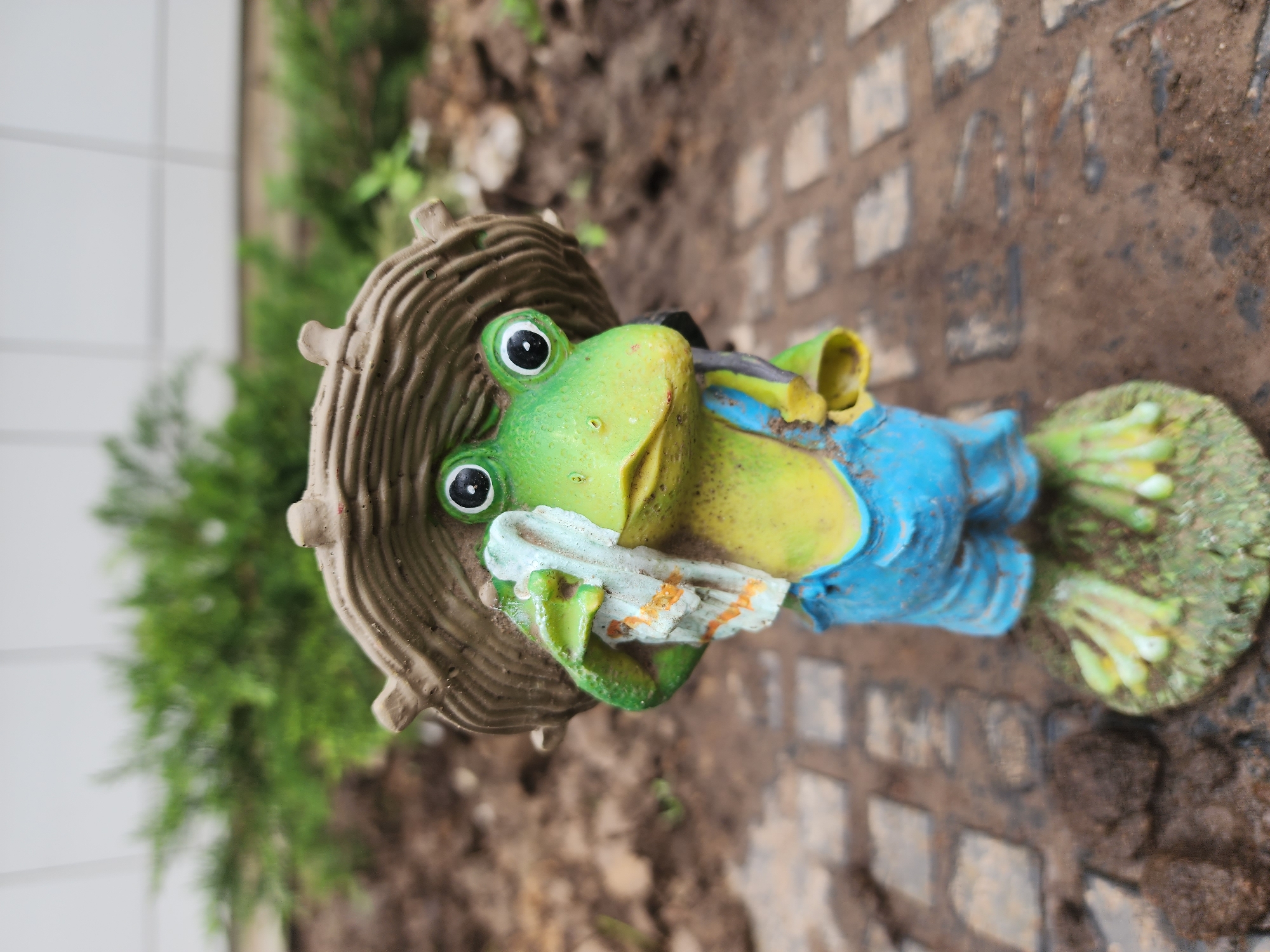 A ceramic frog figurine with a tan mushroom-shaped hat is positioned on the ground in an outdoor setting. The frog is painted green and yellow with blue overalls, and it holds a white towel with orange stripes. The ground is a mix of dirt, small rocks, and cobblestone bricks. Green foliage is visible in the background. Based on the "A14" visible in the cobblestone, the location is likely Kazan, Russia.