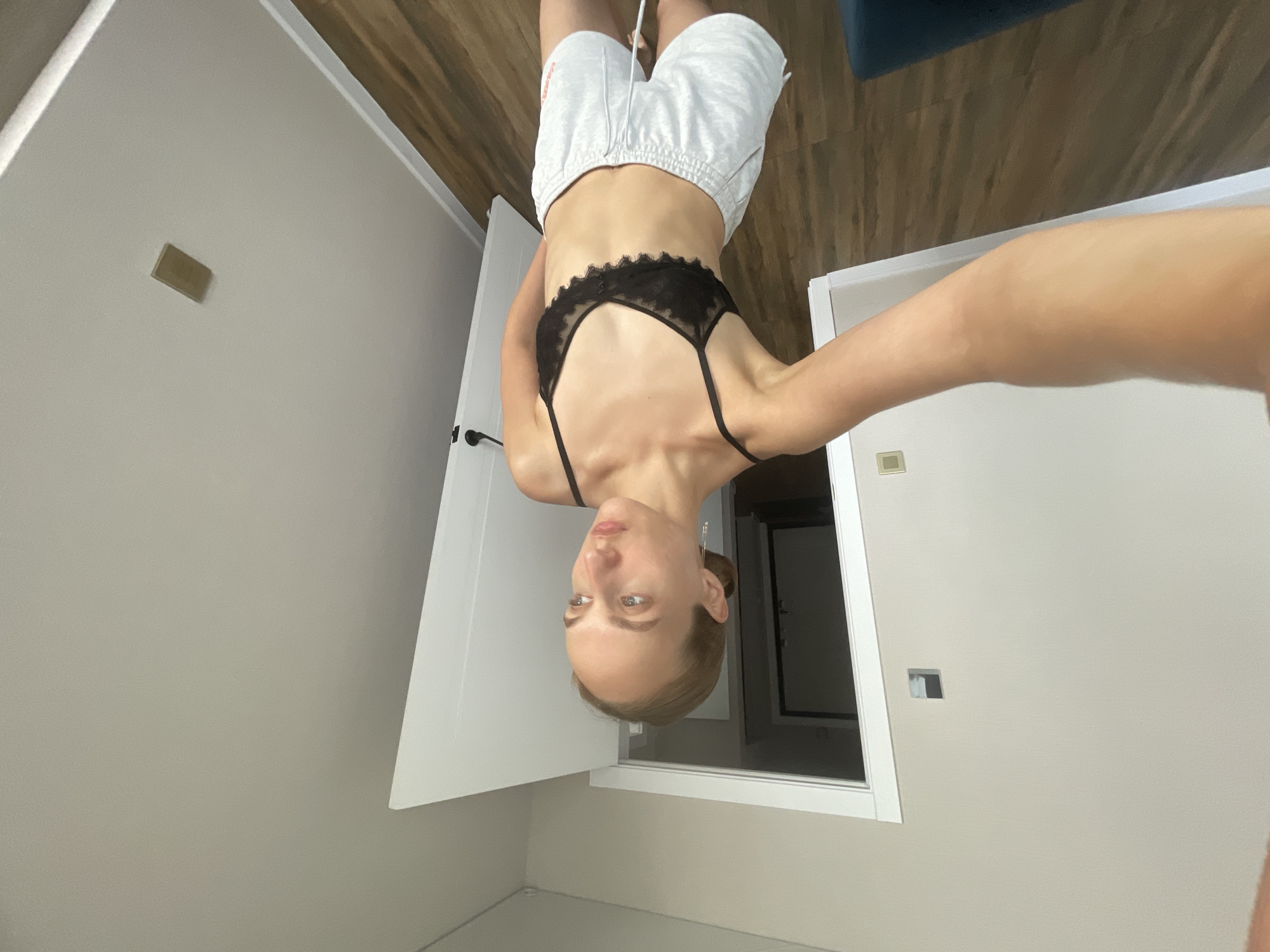 A young woman with blonde hair pulled back stands in a brightly lit room, taking a selfie with her left arm extended. She is wearing a black lace bralette and light grey sweat shorts with an orange logo on the right leg. The room has light beige walls and a wooden floor. To her left, a doorway leads to another room with a darker interior and a closed door. To her right, a white door is ajar. A light switch or outlet is visible on the wall behind her, and another rectangular object is mounted on the wall to the right. The overall scene suggests a casual, domestic environment. The location is identified as Podshivalovo, Russia.