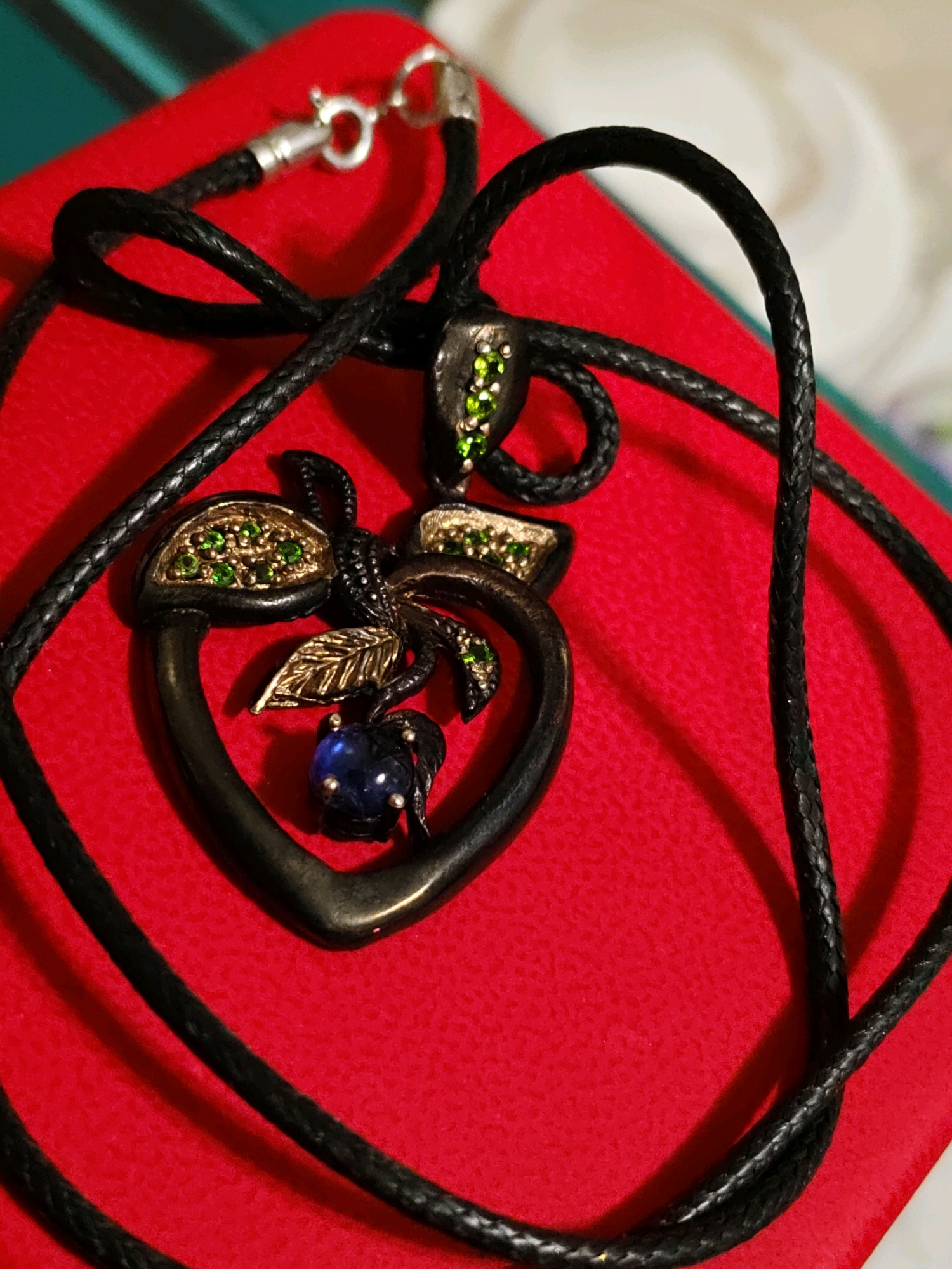 A black cord necklace with a heart-shaped pendant is displayed on a textured red surface. The pendant is dark in color, possibly metal, and is adorned with green gemstones and a single blue gemstone at its base. A small gold-colored leaf detail is also present on the pendant. The necklace has a silver clasp. The background appears to be a deep green surface with some subtle lighting effects. The image is taken from a top-down perspective, focusing on the necklace. There are no people or specific locations in the city of Rostov-on-Don, Russia, identifiable in the image.
