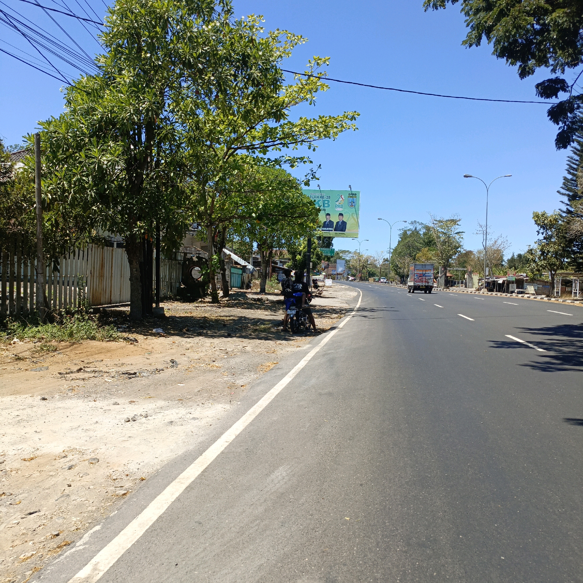 The image depicts a street scene in Salatiga, Indonesia. A paved road runs from the foreground into the distance, bordered by a sandy shoulder and vegetation on the left. Trees line the roadside, with a wooden fence visible behind them. A billboard featuring two men in suits is positioned prominently on the left side of the road, near the trees. A motorcycle with two people seated on it is parked on the shoulder, under the shade of a tree, next to the billboard. Further down the road, a truck and other vehicles are visible, indicating traffic. The sky is clear and blue, suggesting a sunny day. The overall environment appears to be a semi-urban or roadside setting within Salatiga.