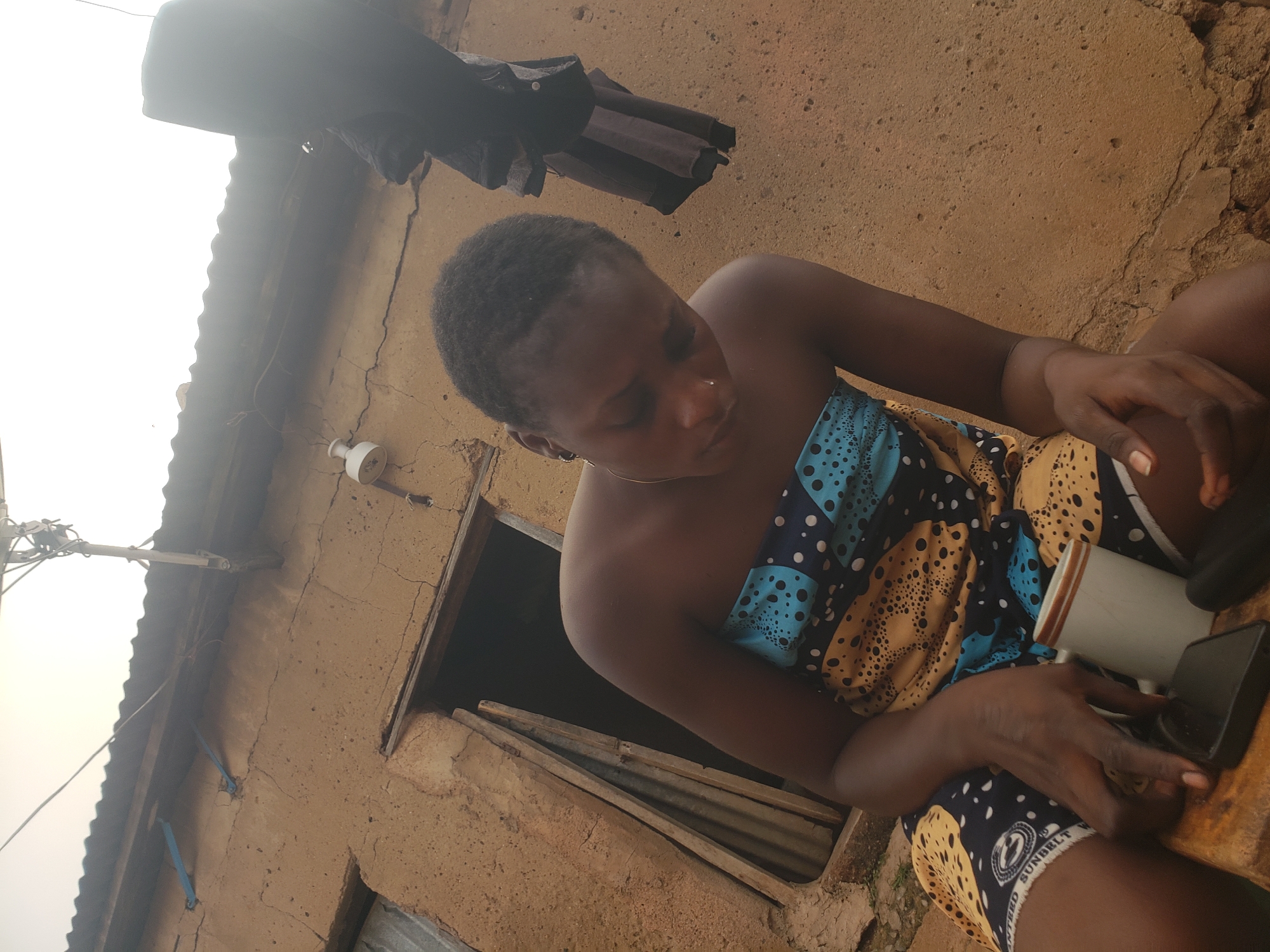 A medium, angled shot depicts a woman holding a smartphone and a white mug in her lap, seated outside of a building in Bukuru, Nigeria. The woman, of African descent, has short dark hair and wears a colorful patterned dress. Her head is tilted down as she focuses on the phone in her hands.
The background reveals an exterior wall of a building with a doorway or window. Above her hangs what appears to be clothing or fabric, drying on a line. Electrical wires and a light fixture are attached to the building's exterior. The building appears to be made of clay or earth.