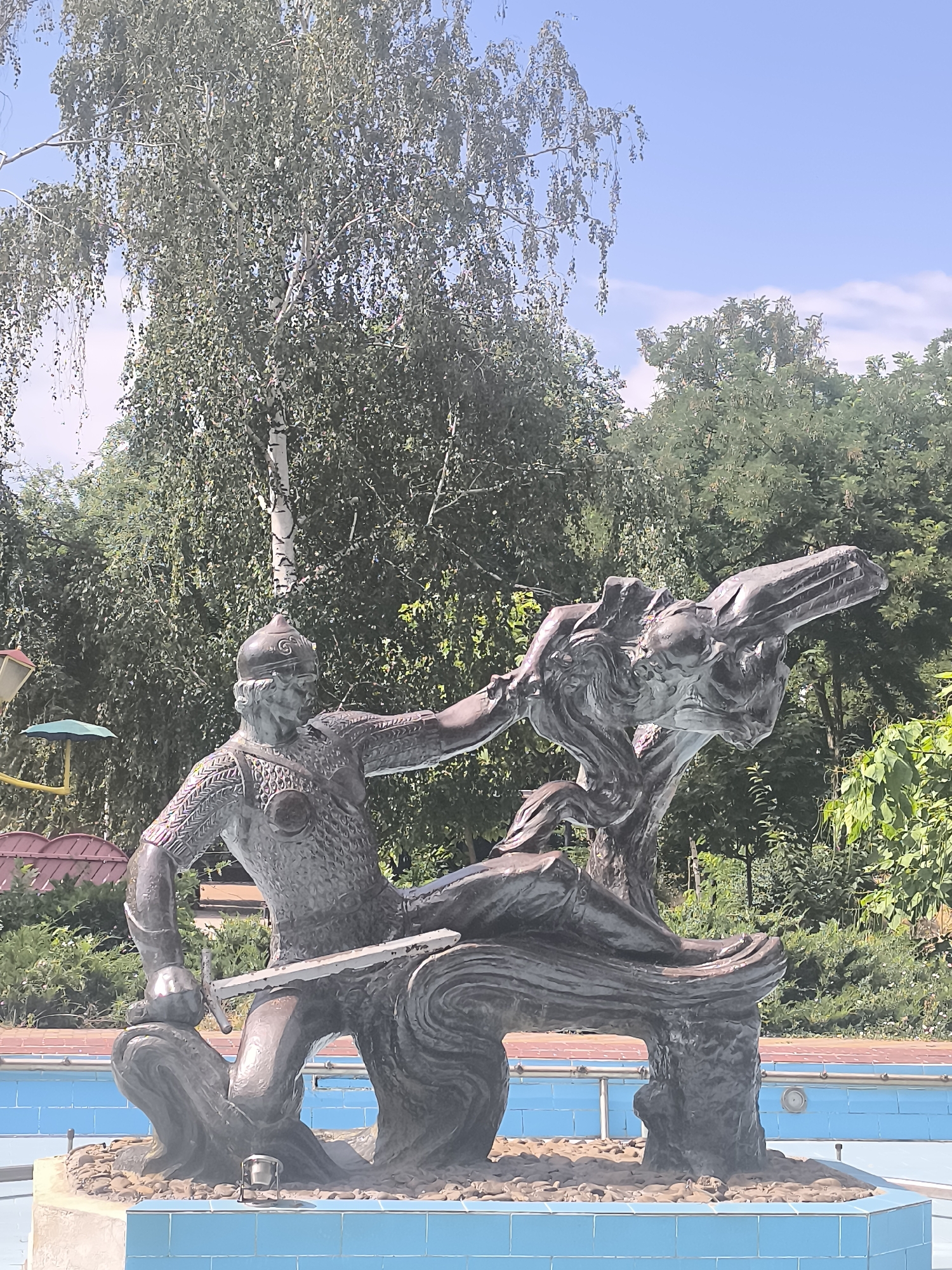 This is a medium shot of a bronze statue of a warrior fighting a serpent, located in Dnestrovsk, Moldova. The statue depicts a man in ornate armor, holding a sword, and seemingly engaged in a struggle with a dragon-like creature. The warrior is seated on a stylized, wave-like base, with his legs crossed and his upper body leaning forward. He is wearing a helmet, a patterned tunic, and armored leggings. His right arm is extended, gripping the serpent's head, while his left hand holds the sword which is positioned across his lap. The serpent is coiled around a tree-like structure that forms part of the base, with its head raised and its mouth open. The statue is positioned next to a swimming pool, the blue tiled edge of which is visible in the foreground. Behind the statue, lush green trees and foliage form a backdrop, with a clear blue sky overhead. The overall impression is one of a mythological or historical scene, rendered in a dynamic and expressive style.