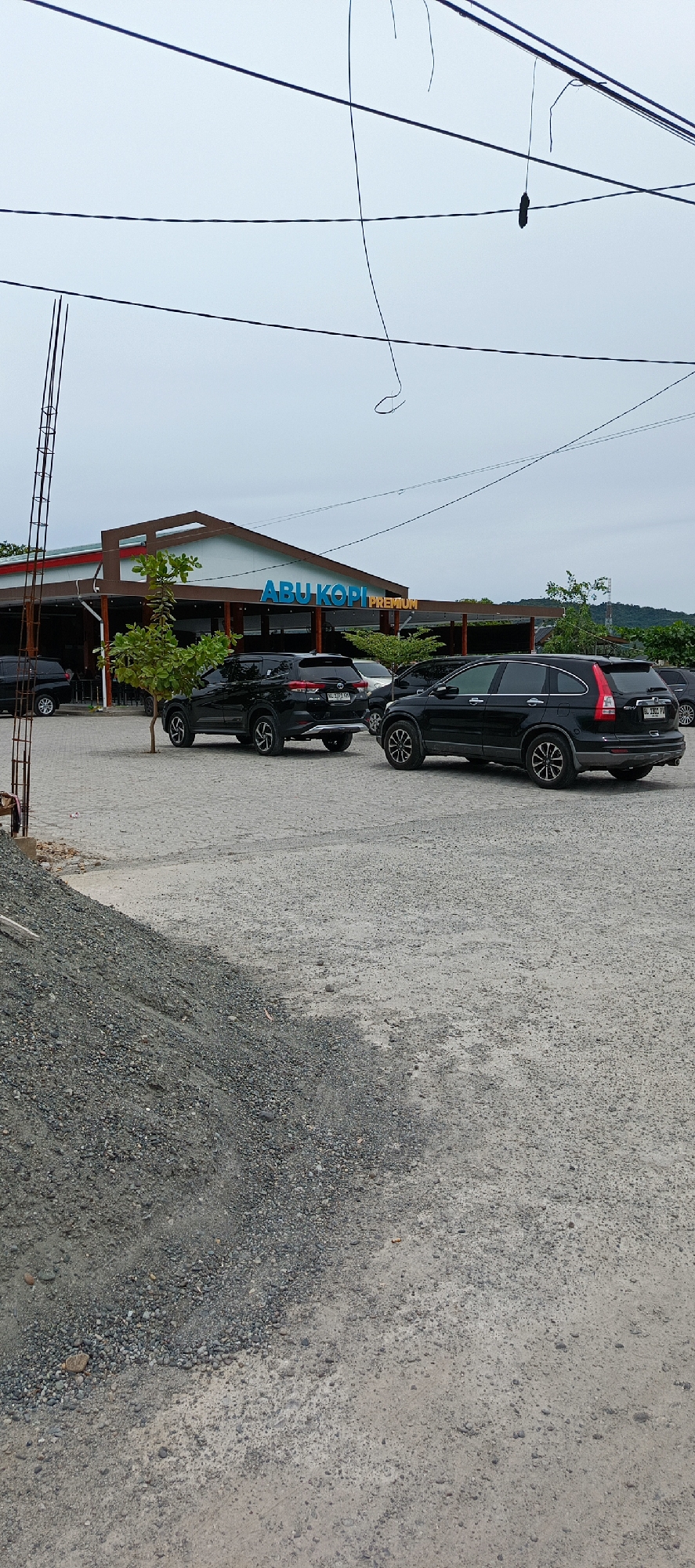 The image depicts an outdoor scene in front of a building with a sign that reads "ABU KOPI PREMIUM" in blue and yellow lettering. The building has a light blue facade and a brown roof. Several black SUVs are parked in a gravel lot in front of the building. A small tree with green leaves stands in the foreground. The sky is overcast and appears to be a light gray color. The ground is covered in gravel and appears to be a parking area. In the background, there is a glimpse of a hillside covered in trees. There are electrical wires visible in the sky above the scene. The overall impression is of a cafe or restaurant setting.