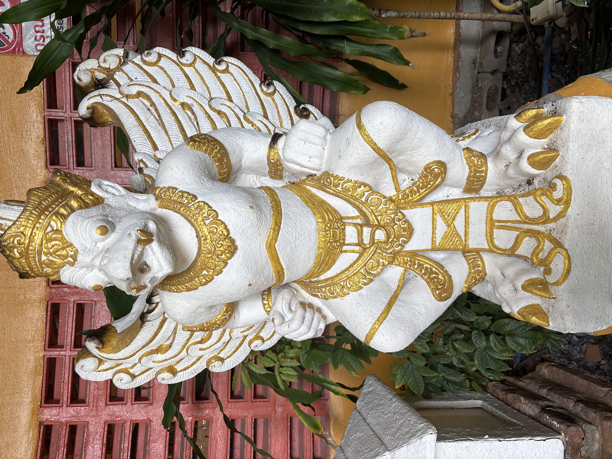 A white statue with gold accents depicts a bird-like mythical creature with large wings, a crown, and sharp talons. The statue is positioned in front of a red brick wall with yellow trim and is surrounded by lush green foliage. A small white structure with a pointed roof is visible to the left of the statue. The overall scene suggests an outdoor setting, possibly a temple or garden, in Chalong, Thailand. The statue is stationary and appears to be an ornamental piece.