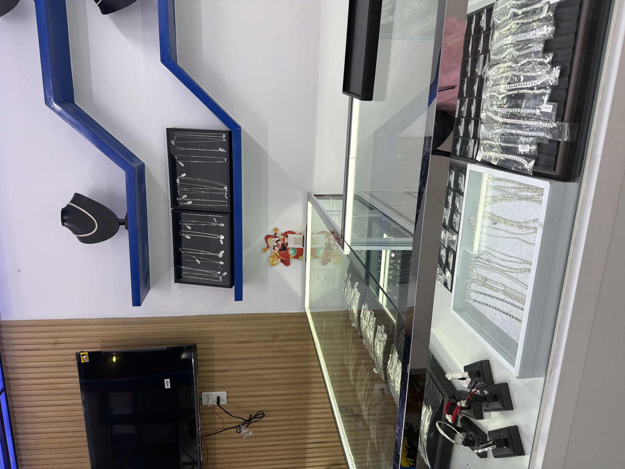 This image captures the interior of a jewellery shop, likely a retail display area, presented with a sideways orientation. The space is brightly lit and appears well-organized.
On the left side of the frame (which would be the back wall if correctly oriented), a white wall is adorned with striking blue, angular shelves. Two black necklace mannequins are mounted on these shelves, each showcasing a sparkling, silver-colored necklace. Below them, two black display trays feature multiple delicate silver-toned necklaces, each individually sealed in a small clear plastic bag.
Further down the left, a section of the wall is covered with horizontal wooden paneling. A black flat-screen television is mounted here, currently off or displaying a dark screen. Below the TV, a white electrical outlet has a charger plugged in, with red and black wires visible on the wooden paneling.
The right side of the image (the foreground) is dominated by various glass display cases. A prominent, brightly lit glass display counter features an LED strip along its edge, illuminating numerous silver-colored bracelets, many individually wrapped in plastic. Adjacent to this, a white display tray presents more silver-toned necklaces or bracelets. Closer to the viewer, several smaller black stands showcase individual pieces of jewellery, including bracelets. One stand displays a red and black string bracelet.
A small, colorful sticker featuring a cartoon figure, resembling a lucky charm or deity often associated with prosperity (like Caishen), is visible on the back wall near the display cases.
Visible text includes "790.00" on a price tag attached to a bagged bracelet within the large glass display case, and "700" on a small tag on one of the individual jewellery stands. No people are present, indicating a static view of the merchandise. The items and display suggest a focus on silver or silver-colored jewellery, potentially reflecting a contemporary style. The clean and organized presentation implies a commercial setting.