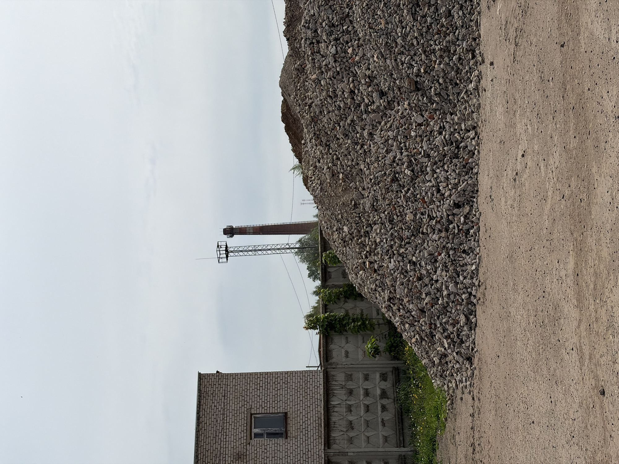 The image depicts an industrial scene in Uva, Russia. A large pile of grey gravel dominates the right side of the frame, alongside a dirt road. In the background, there's a brick building with a single window, a concrete fence with climbing vegetation, and a tall metal tower. A brick smokestack is also visible. The sky is overcast. There are no people visible in the frame.