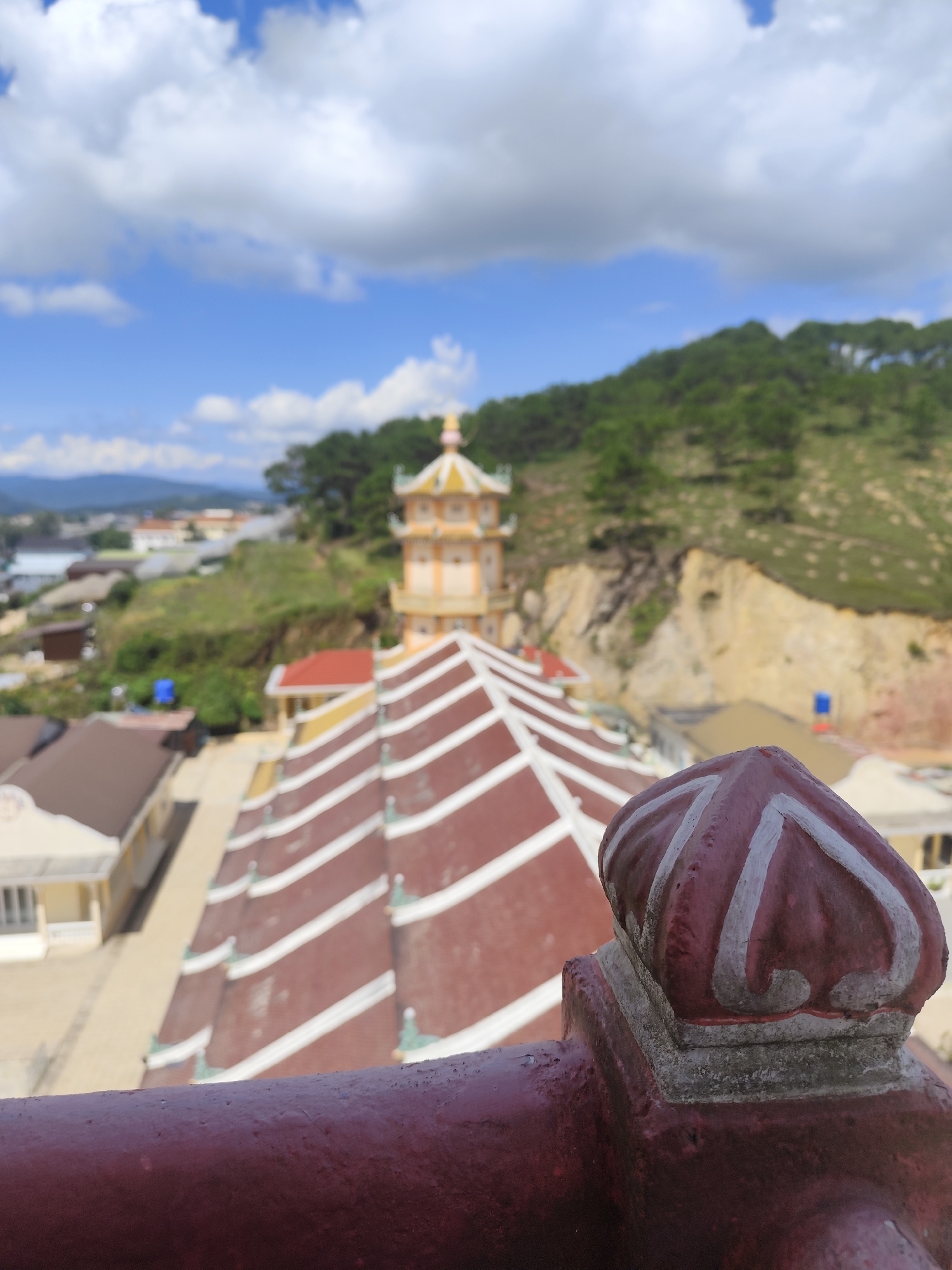 The image is a medium shot, taken from an elevated viewpoint, looking down on a temple complex and its surrounding environment. The foreground is dominated by a dark red railing and a decorative finial, which appears to be made of plaster or concrete and painted with white swirling patterns. The finial is situated on a square base.
Beyond the railing, the focal point is a large, multi-tiered pagoda-like structure with a distinctive red roof adorned with white stripes. The roof has a complex, stepped design. To the left of this structure, several buildings with cream-colored walls and brown roofs are visible.
In the mid-ground, a tall, yellow tower with multiple levels and a decorative yellow cap stands prominently. This tower also features architectural elements characteristic of East Asian temples. The buildings and the tower are situated on a hillside, which is covered with green trees and vegetation. The terrain appears to be uneven, with some exposed soil and rock faces visible.
The background reveals a hazy cityscape with more buildings and infrastructure stretching into the distance. Further beyond the city, a range of green mountains forms the horizon. The sky above is a vibrant blue, punctuated by fluffy white clouds.
The image appears to be taken in Lộc Quý, Vietnam, given the architectural style of the temple complex and the hilly, vegetated landscape often found in the region. There are no people visible in the image. The overall scene depicts a tranquil temple setting within a broader urban and natural landscape. The lighting suggests it is daytime, possibly late morning or afternoon, given the bright illumination and clear sky.