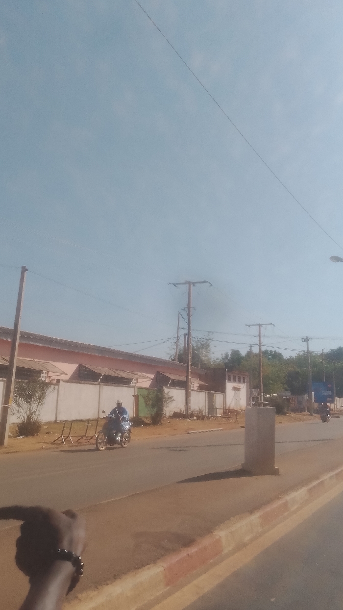 The image depicts a daytime street scene in Korhogo, Côte d'Ivoire. The perspective is from an individual standing by the roadside. In the immediate foreground, a dark-skinned hand with a dark beaded bracelet is visible in the bottom-left, pointing forward. The scene features a two-lane asphalt road with a red and white painted curb delineating a median or sidewalk on the right. A grey concrete pillar stands on this median.
On the left side of the road, a light-colored building with a dark, sloped roof and several window-like openings along its upper section is set back behind a white concrete perimeter wall. Scattered green foliage is present between the wall and the building, as well as along the roadside. A tall concrete utility pole with overhead wires stands prominently on the left.
Mid-ground, a person wearing a light-colored helmet and blue clothing is operating a light-colored motorcycle in the left lane, traveling from left to right across the frame. Further down the road on the right, a second, less distinct motorcyclist is visible. Multiple utility poles of various materials (concrete, wood) carrying overhead electrical or communication lines extend along both sides of the road. The sky is clear and light blue. In the distant background on the right, additional trees, buildings, and a streetlamp are discernible.