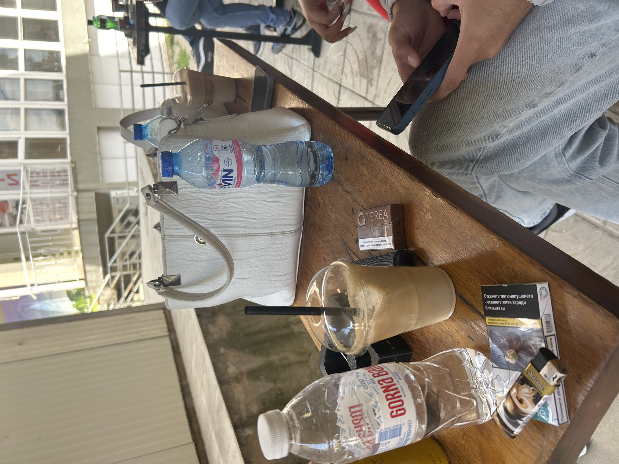 The scene depicts a wooden table, likely outdoors, with various items scattered across it. A fair-skinned individual wearing light-colored denim jeans is holding a smartphone, focusing on the screen. On the table, there's a white handbag, two bottles of water (one labeled "MЯN" and another "GORNA BANYA"), a plastic cup filled with a coffee-like beverage and a black straw, a packet of "TEREA TEAK" cigarettes, and a pack of cigarettes with a lighter on top. The text on the cigarette pack reads "Откажете тютюнопушенето- останете живи заради близките си Помощ за отказ от тютюнопушене: 0700 10 323" which translates to "Quit smoking - stay alive for your loved ones. Help to quit smoking: 0700 10 323." In the background, there is a blurred view of a building with windows and other seated individuals. Based on the brands of the water and cigarette packets, and the language of the cigarette warning, the location is likely Blagoevgrad, Bulgaria.