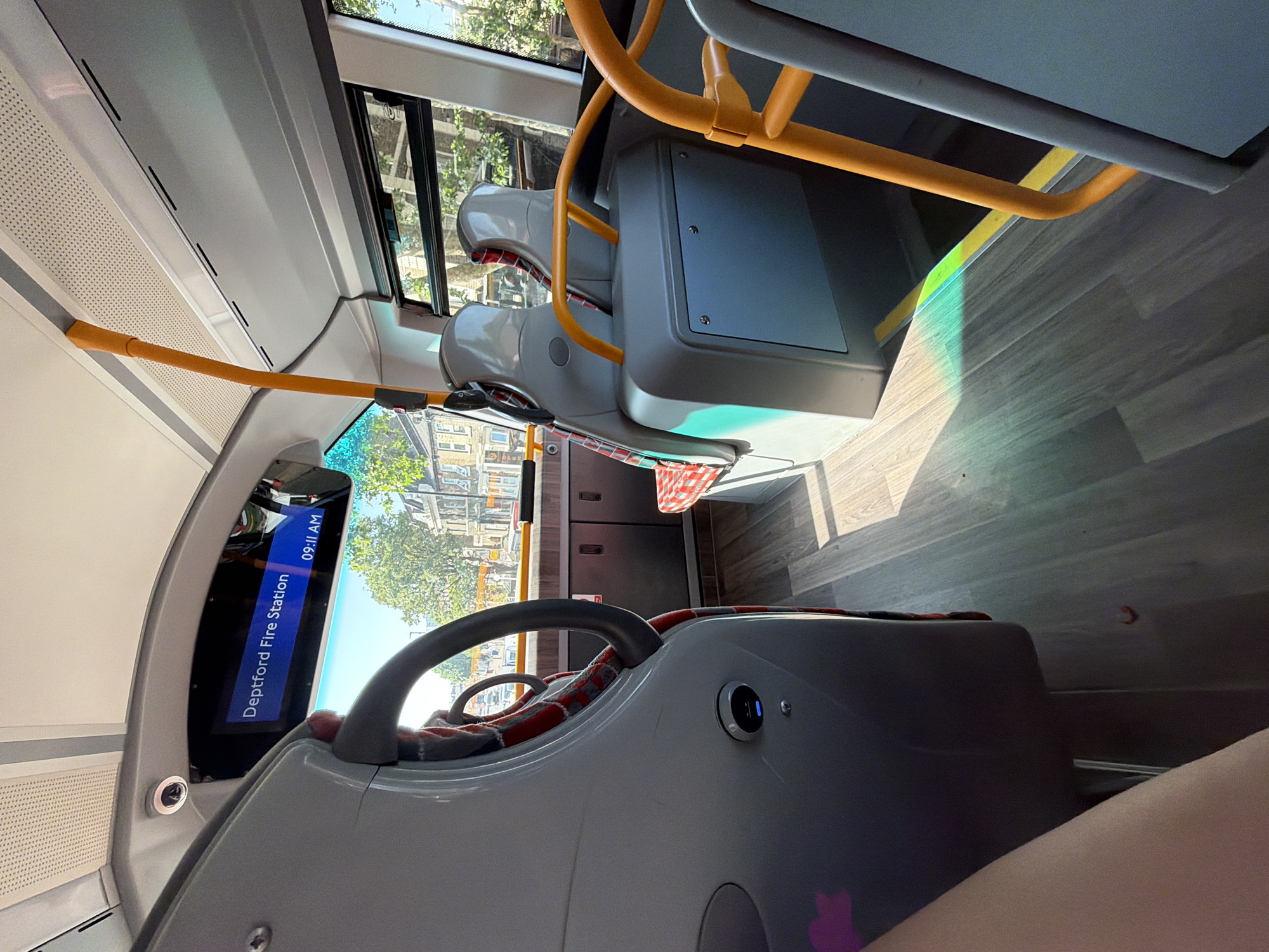 The image shows the interior of a bus in London, United Kingdom. The destination display reads "Deptford Fire Station" and the time is 09:11 AM. The bus has grey seats with red patterned fabric. There are yellow handrails. Outside the window, buildings and trees are visible. The floor is made of wood-effect planks. A portion of a person's leg is visible in the foreground.