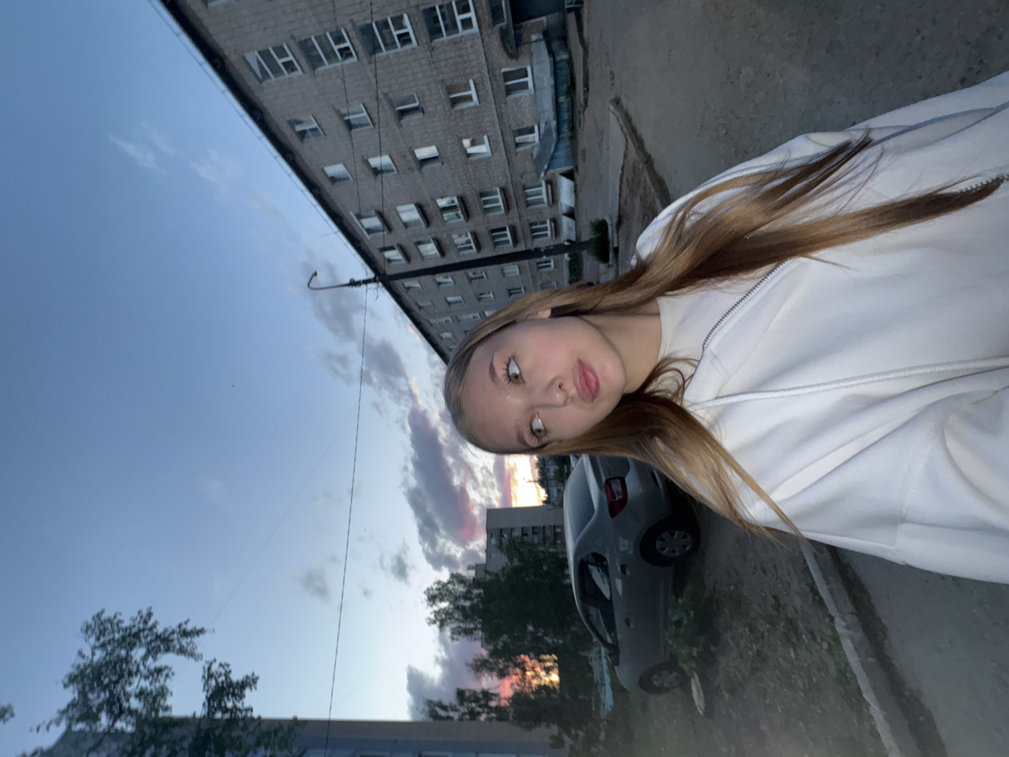 The image shows a young woman in a white hooded sweatshirt, standing on a sidewalk next to a gray car. Behind her are apartment buildings, trees, and a sunset sky. The buildings have multiple windows and a gray facade. The car is parked on the side of the road. The location appears to be in a residential area of a city. The composition suggests the photo was taken in an urban setting, possibly in Chaikovsky, Russia, based on the building architecture and general environment.