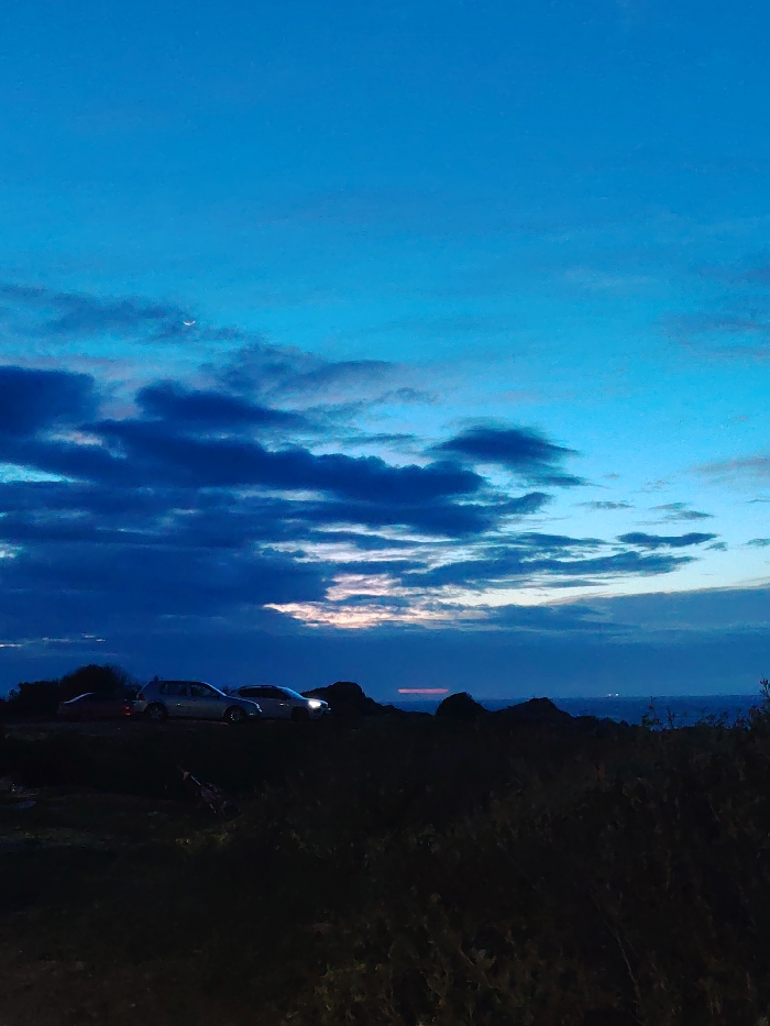 This image depicts a coastal twilight scene, likely at a high vantage point overlooking the sea near Tanger, Morocco. The sky occupies the upper two-thirds, exhibiting a gradient from a deep indigo at the horizon to a lighter cerulean blue at the zenith. Scattered dark cumulus clouds are prominent across the middle band of the sky, with a sliver of a crescent moon visible in the upper left. A faint, reddish-orange glow from the setting sun is discernible beneath the clouds on the distant horizon line.
The immediate foreground is composed of dark, indistinct vegetation and uneven terrain. In the midground, three vehicles are parked on what appears to be a dirt or gravel surface. From left to right, a partially visible red vehicle is parked, followed by a silver-colored Volkswagen Golf hatchback, and then a white SUV, possibly with its headlights emitting a faint glow. A darker, less distinct vehicle is positioned between the Golf and the SUV. No individuals are visible.
Beyond the vehicles and the dark landmass, a wide expanse of dark water, presumably the Atlantic Ocean or the Strait of Gibraltar, extends towards the horizon. Faint, scattered lights of a distant city or coastline are visible on the far horizon, approximately in the center to right of the frame. A distinct, horizontal reddish light streak is observable above the distant water on the right side of the image.