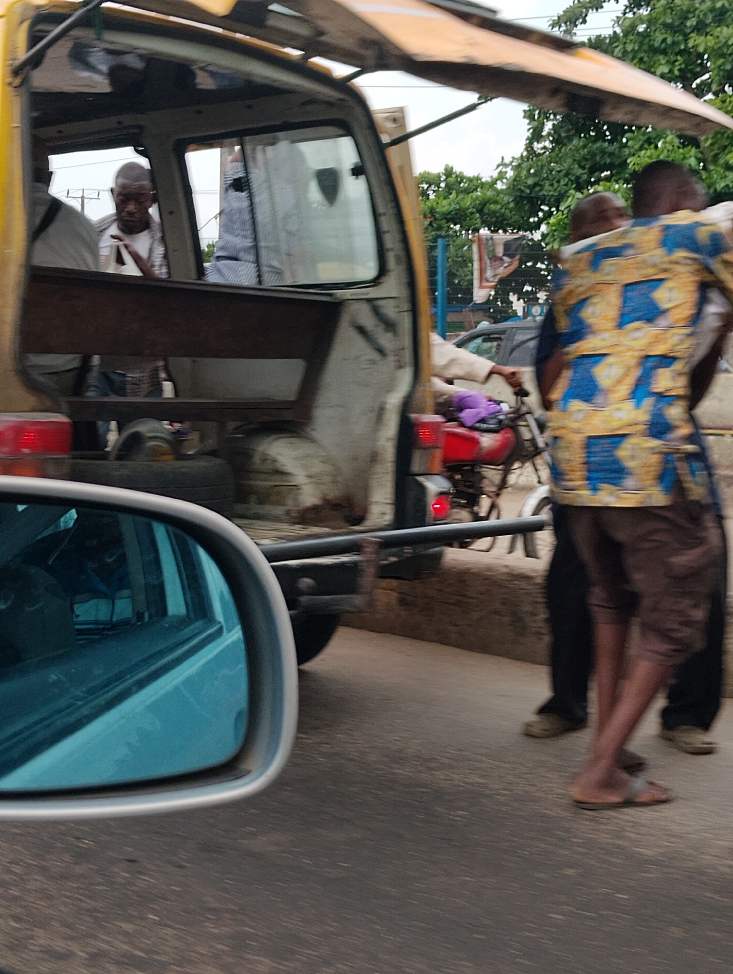 In Shangisha, Nigeria, a yellow vehicle, possibly a repurposed van, is parked by the roadside. Through its open rear, we see a man wearing a white shirt and what appears to be a blue and white striped garment. He is holding a white piece of paper and looking downwards. Another person, mostly obscured, is seated beside him. The vehicle has a wooden bench inside and what looks like spare tires and a spare wheel are visible in the rear cargo area.
Outside the vehicle, a person wearing a vibrant blue and yellow patterned shirt and brown shorts is standing. Their legs are crossed, and they are facing away from the camera. To their left, a red motorcycle is parked. A purple cloth is draped over the back of the motorcycle seat. Another person, in dark trousers and light-colored shoes, stands behind the person in the patterned shirt.
The scene appears to be set during daylight, with lush green trees visible in the background, suggesting a warm or humid climate. The road surface is asphalt. A side mirror of a car, possibly the one from which the photo is taken, is in the foreground, reflecting a blue sky and some greenery. The overall impression is a snapshot of everyday street life in a Nigerian town. There is no visible text in the image.
