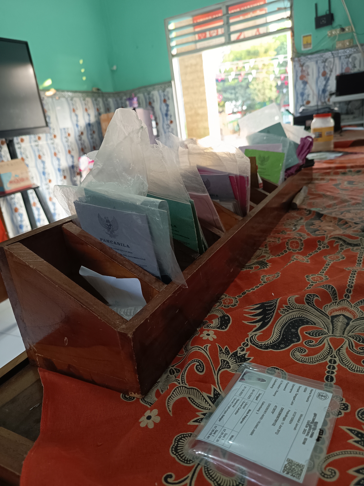The image shows a wooden desk organizer filled with various documents, most of which are enclosed in clear plastic sleeves. One document in the foreground of the organizer clearly displays the text "PANCASILA" and a Garuda bird emblem. The organizer is placed on a table covered with a red and black patterned cloth, possibly batik. In the background, a window with horizontal blinds allows sunlight to enter, illuminating a room with turquoise walls and a tiled floor. A computer monitor is visible on the left, and a shelf with what appear to be some sort of jars and other items is on the right. The scene appears to be indoors, likely in a residential or office setting in Widoro, Indonesia, given the context of the "PANCASILA" document, which is the state ideology of Indonesia. No people are present in the image.