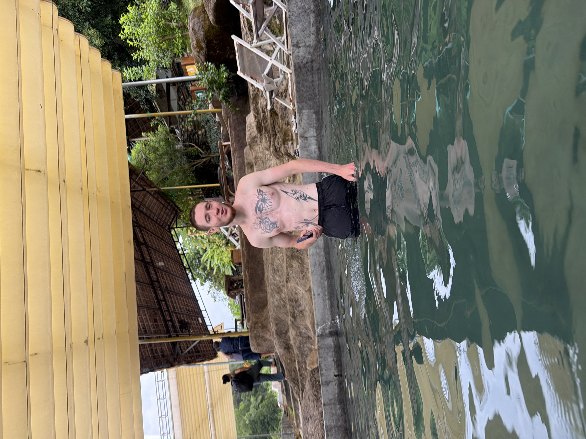 A young Caucasian male with tattoos on his chest and abdomen stands in the water of a pool, wearing black shorts. He is holding a dark-colored smartphone in his right hand. The man has short, light brown hair and a beard. In the background, two other individuals are walking up a series of stone steps. One is wearing a dark backpack, and the other is wearing a dark blue shirt. Further in the background, there is lush green foliage, two lounge chairs, and a thatched-roof structure. The scene appears to be an outdoor resort or natural hot spring area in Khanh Vinh, Vietnam. The water is clear and reflects the surrounding environment.