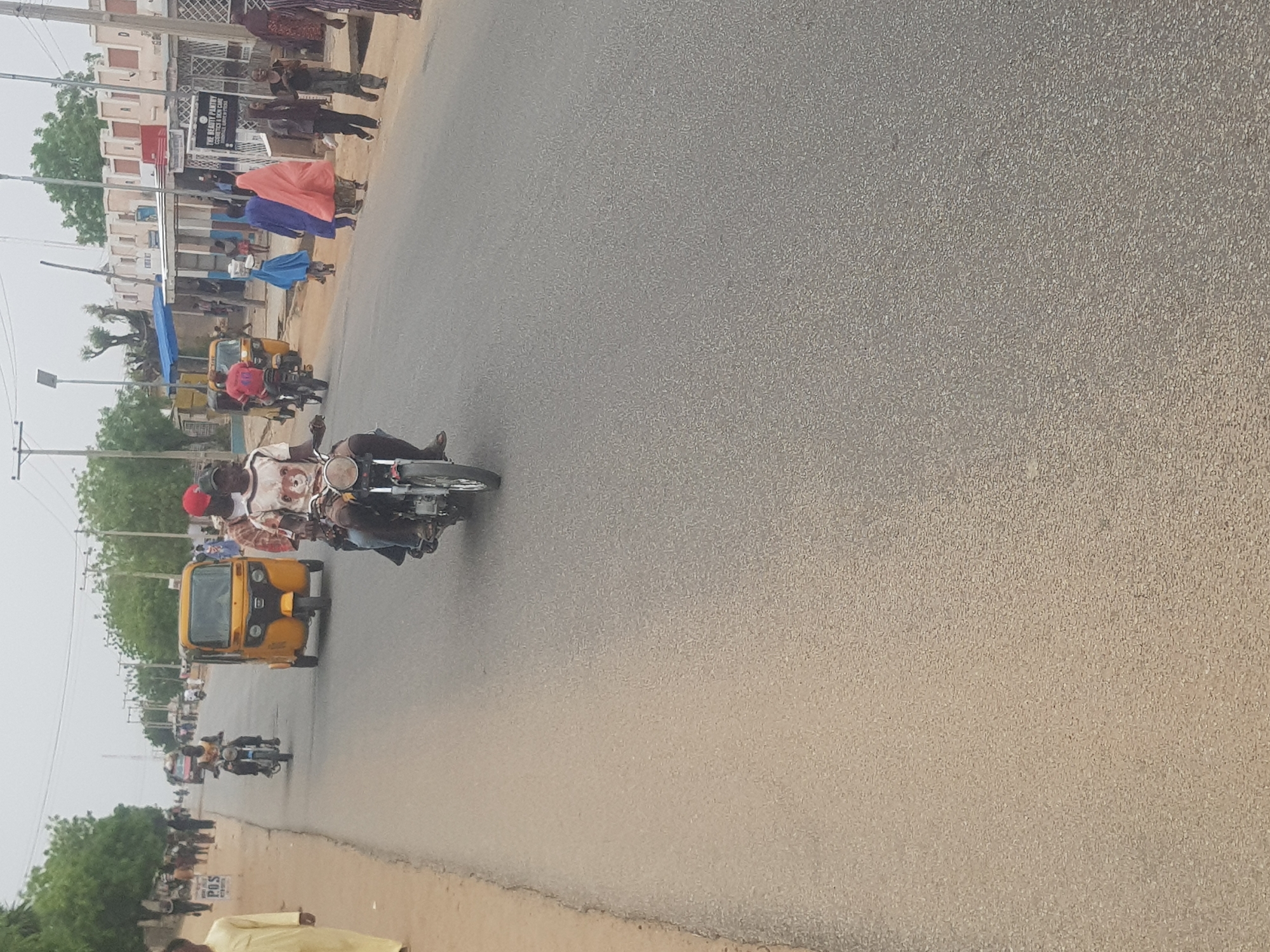 The image depicts a street scene in Dabawa, Nigeria. The road, which takes up most of the frame, is paved and appears to be made of asphalt. A motorcycle with two riders, one wearing a red hat, is traveling along the road. In the background, there's a yellow tricycle and another motorcycle, also in motion. On the left side of the road, a group of people are walking on a sidewalk in front of buildings. Some are wearing brightly colored clothing, possibly robes or wraps. Trees and utility poles are visible alongside the buildings. The sky is overcast.
