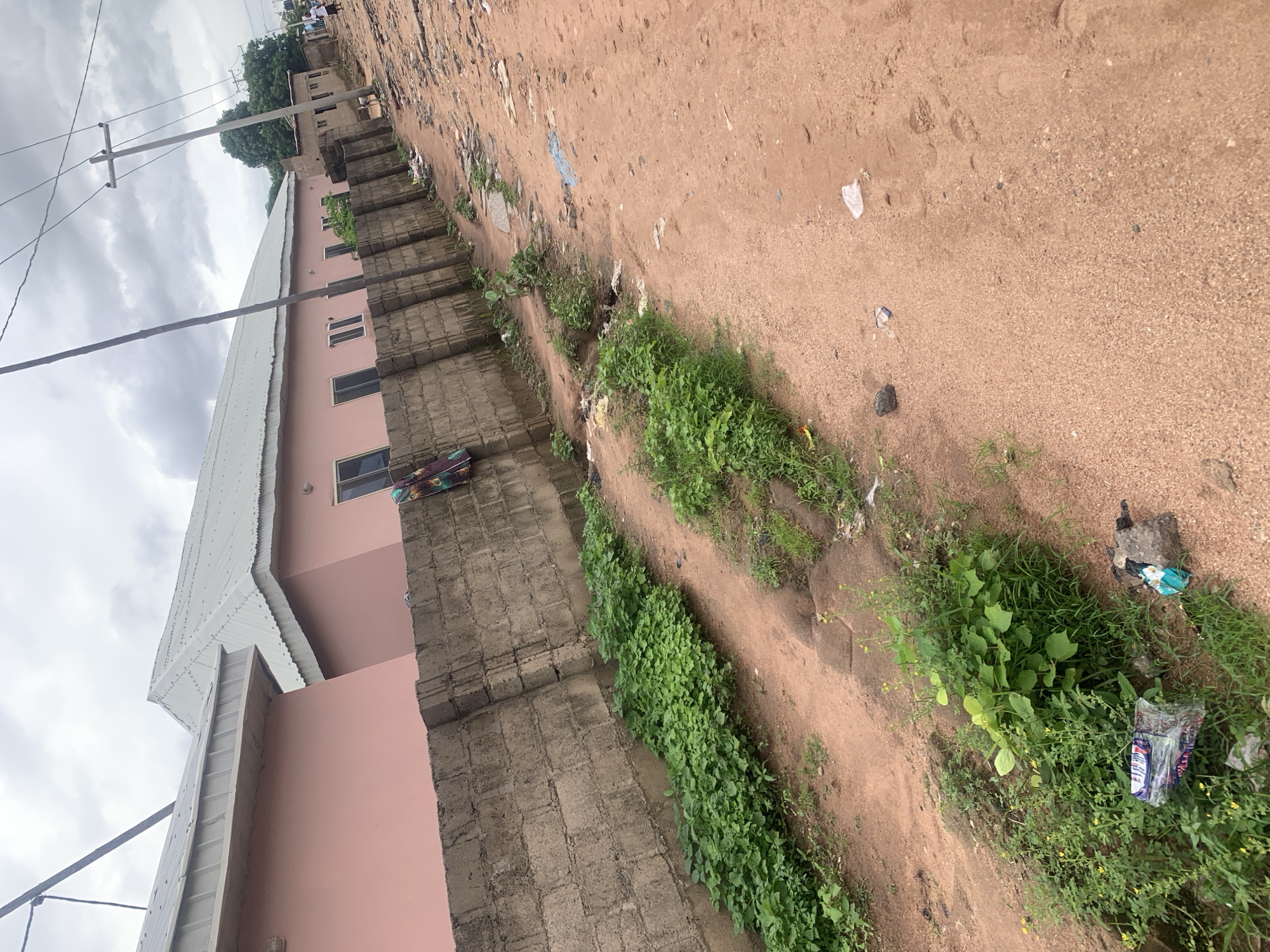 The image depicts a dirt road in Jalingo, Nigeria, with buildings and vegetation on either side. On the left, there are two pink buildings with white corrugated metal roofs, separated by a concrete block wall. The buildings have multiple windows and a few sparse trees are visible behind the wall. The wall itself is covered in some areas by lush green vegetation. The road in front of the buildings is a sandy dirt track, with some litter scattered along its edges. In the foreground, to the left, is a pile of plastic packaging and a small pile of debris. Further down the road, to the right, the terrain becomes more uneven with more debris visible. Electrical poles and wires are present in the background, and a few people can be seen walking in the distance on the right side of the image. The sky is overcast.