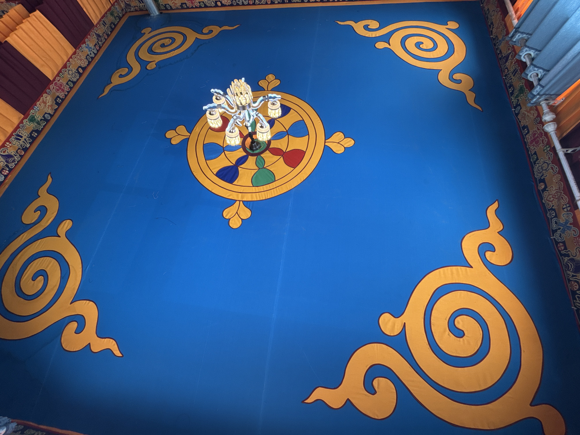 The image shows the ceiling of a building, possibly a temple, in Jila, China. The ceiling is painted blue and adorned with stylized gold swirl patterns in each corner. In the center is a large gold Dharma wheel design with red, green, and blue sections. A modern chandelier is suspended in the center of the Dharma wheel. The edges of the ceiling are decorated with patterned borders of yellow, red, and maroon. The overall impression is that of an ornate, possibly sacred, space.