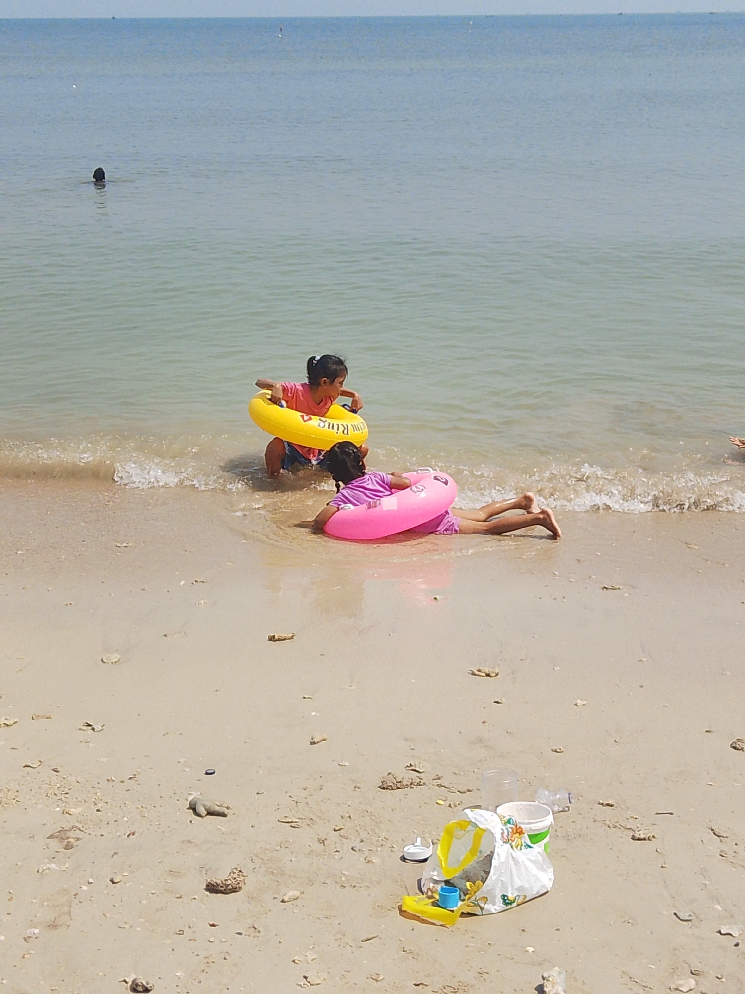 This media file depicts a serene beach scene with two young girls enjoying the water. The setting is a sandy beach, with the gentle waves of the ocean lapping at the shore. The sky is clear and bright, suggesting a sunny day, likely in the late morning or early afternoon.
One girl, wearing a pink shirt and shorts, is lying on her stomach in the shallow water, partially submerged and with a pink inflatable ring around her waist. She is positioned further from the shore, with her legs extended towards the ocean. Her posture suggests a relaxed or playful engagement with the water.
Another girl, in a pink shirt and blue shorts, is closer to the shore, also with an inflatable ring, this one yellow, around her. She is in a more upright position, appearing to be interacting with the ring or the waves. Both girls have their hair tied back, practical for a beach setting.
In the background, the vast expanse of the ocean stretches towards the horizon, with faint hints of what might be boats or distant landmasses. A solitary figure is visible in the water further out, suggesting other people are also enjoying the sea.
On the sandy shore in the foreground, various objects are scattered, including a white plastic bag filled with what appears to be beach toys like a yellow shovel and blue bucket, along with some discarded plastic bottles and containers. Some small pieces of coral or shells are also visible on the sand. The presence of these items suggests a day of recreation and possibly a clean-up effort or the aftermath of a beach outing.
The overall mood is one of peaceful childhood enjoyment of a natural environment. The location context points to Pejagan, Indonesia, and the image captures a typical beach day. No specific text is clearly legible, apart from what might be "Swim Ring" on the yellow inflatable.