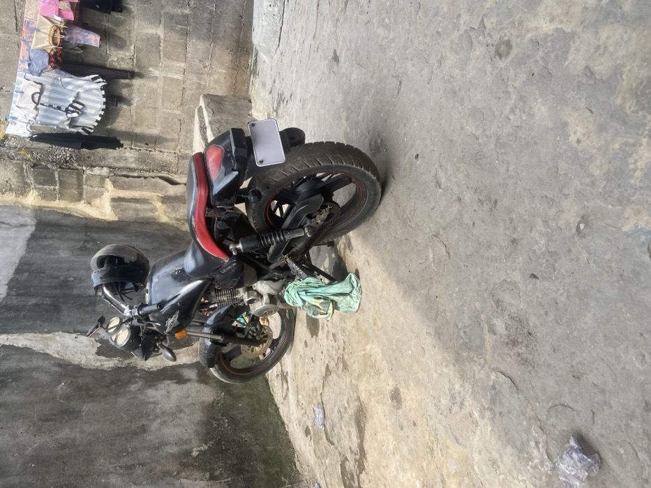 The scene depicts an outdoor concrete area within Alaiyabiagba, Nigeria. A black HOVEN brand motorcycle with a red seat and black rims with red accents is parked centrally on an uneven, stained concrete surface. A black helmet rests on its handlebars, and a light green cloth is draped over the lower engine assembly on the left side. The motorcycle's rear license plate is blank white. To the left, a weathered concrete block wall displays dark discoloration. To the right, another concrete block wall features a clothesline from which various garments, including striped shirts and other colored items, are suspended. A crumpled piece of blue material lies on the concrete ground in the lower-left foreground. No individuals are visible.