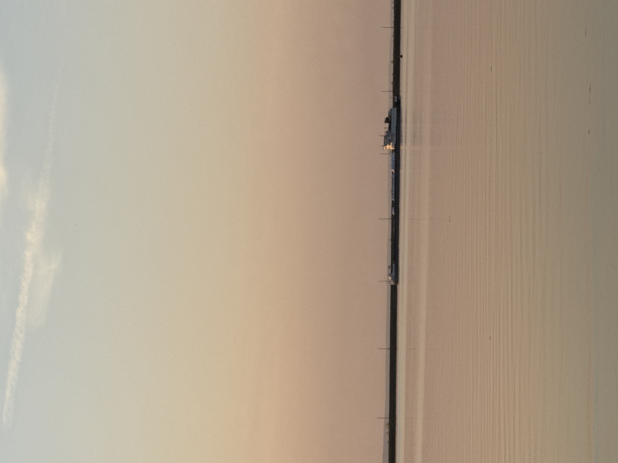 The image depicts the Votkinsk Hydroelectric Station on the Kama River, near Chaikovsky, Russia. The photograph appears to be taken at dawn or dusk. The sky is pale with light clouds on the left. On the right, the calm river reflects the light. A long structure, the hydroelectric station, is situated between the sky and the river.