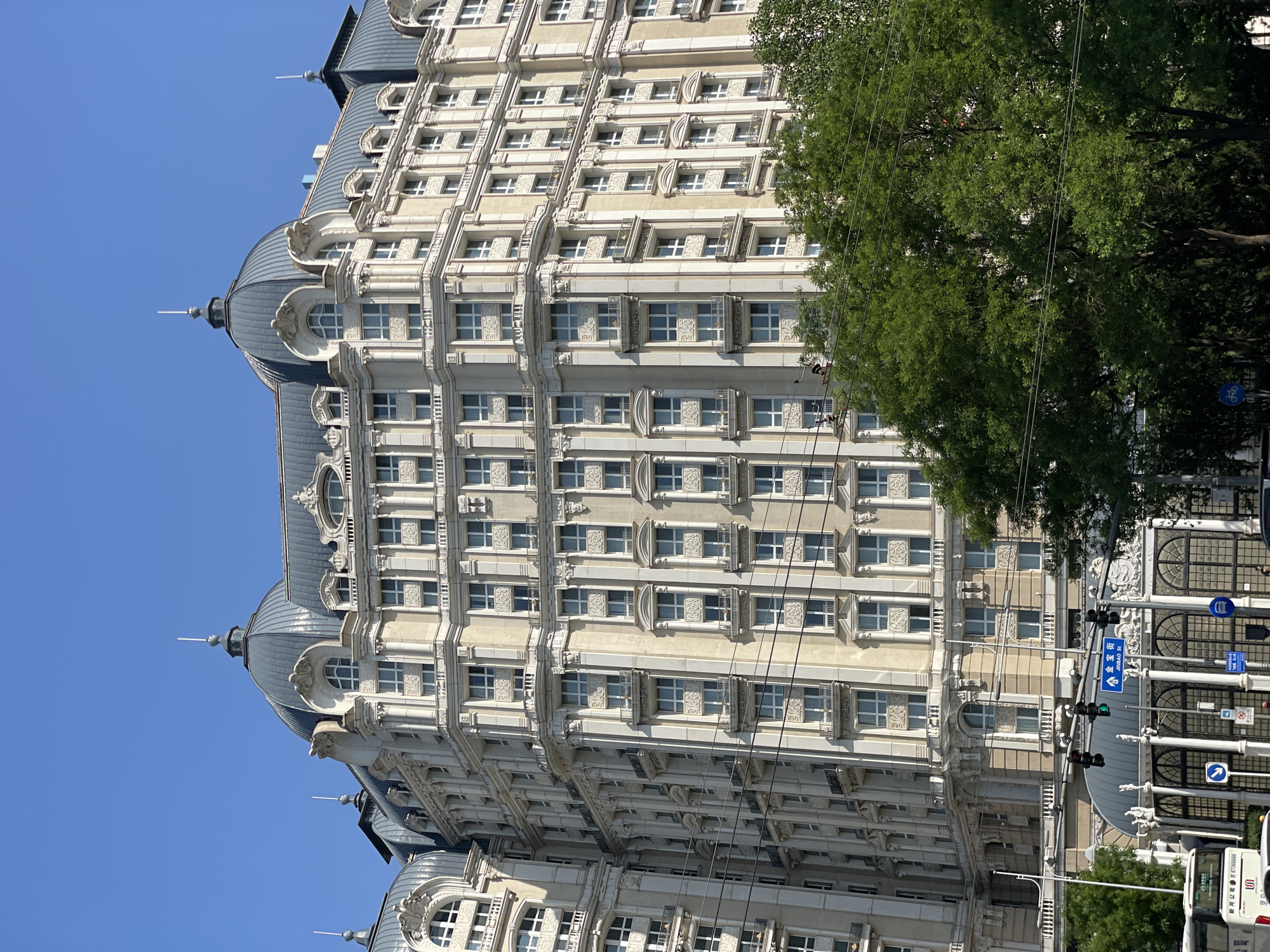 A vertical shot of the Peninsula Beijing Hotel, located in the Dongcheng District of China. The light-colored stone building is ornate in style, featuring rows of windows with white frames, and intricate stonework. At the top of the building are domed roofs. A large green tree obscures part of the right side of the building. In front of the hotel is a sidewalk with a traffic light and street signs. A vehicle can be seen on the far right side of the image. The sky is clear blue.