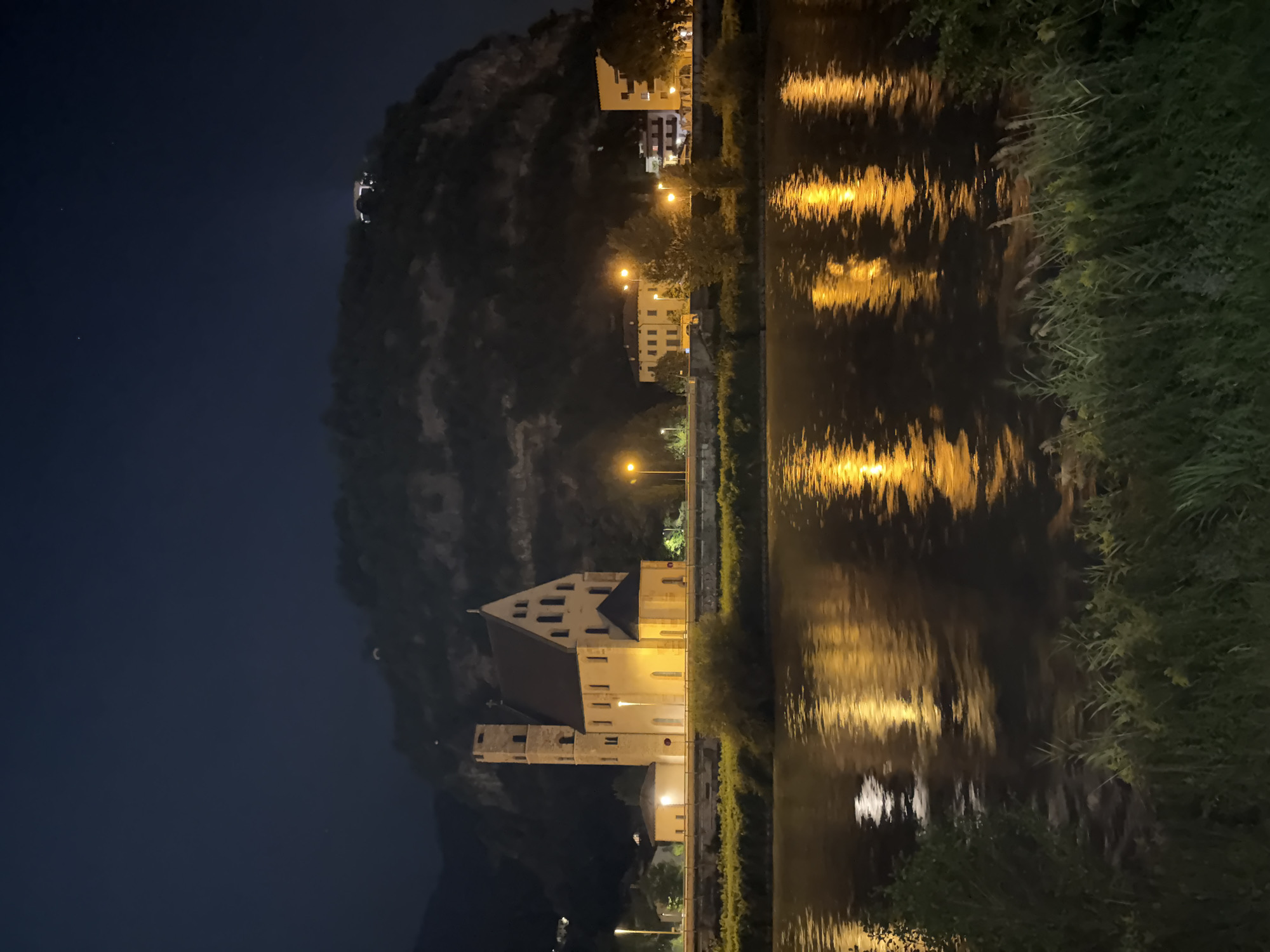 This is a night photograph of the Chiesa di San Vigilio (Church of Saint Vigilius) in Trento, Italy. The church is illuminated and stands prominently on the left side of the frame. It features a tall, square bell tower and a pitched roof. In front of the church, a wide river, likely the Adige River, flows, reflecting the lights from the church and nearby buildings. The water is dark, with shimmering yellow and white reflections.
Behind the church, a steep, rocky hillside rises into the dark night sky. Several buildings, illuminated by streetlights, are visible along the riverbank and on the lower slopes of the hill. The overall atmosphere is serene and picturesque, with the man-made structures contrasting with the natural landscape. The foreground is lined with lush green grass and some reeds. There are no people visible in the image. The sky is a deep, dark blue, with a few distant lights visible on the hillside.