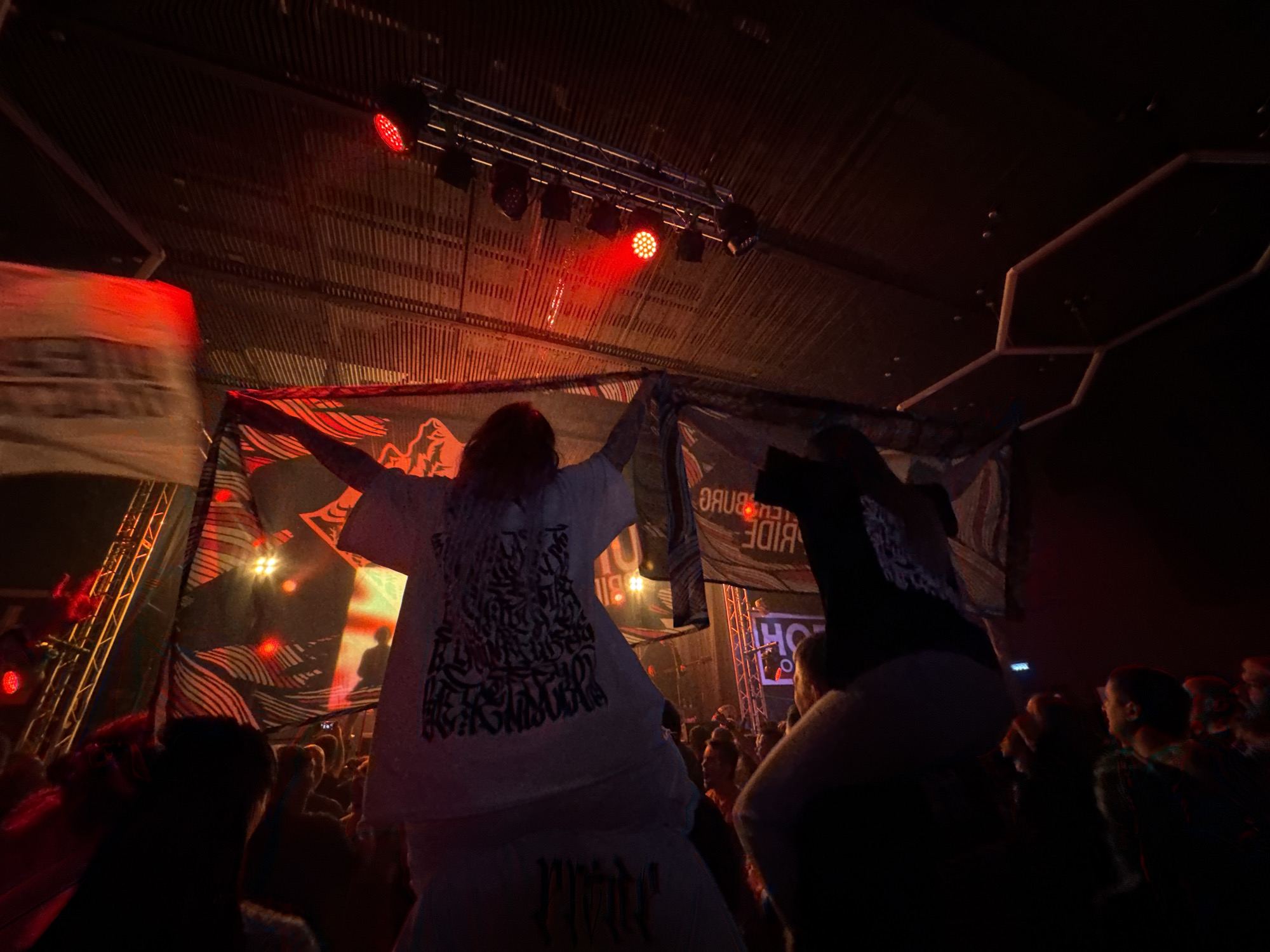 The image depicts a crowded indoor event in Izhevsk, Russia, likely a concert or festival. Several people are visible in the foreground, with one person on someone's shoulders holding a banner with text "Petersburg Pride" displayed backwards. Another individual is holding a banner with text and graphics. Overhead, stage lighting is visible, and the ceiling has a structural pattern. Red stage lights illuminate the crowd.