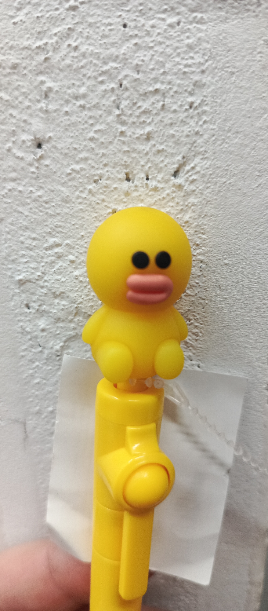 The image shows a close-up of a yellow plastic duck toy with black eyes and a pink mouth, attached to a yellow pen. The duck is positioned centrally in the frame, against a textured white wall that appears to be painted. The wall has a rough, stucco-like finish with small indentations and some darker speckles, possibly due to age or dirt. Below the duck, a portion of the yellow pen is visible, with a white paper label partially attached to its side. The bottom left corner of the image shows a person's hand with light skin, holding the pen. The lighting is even, suggesting an indoor setting, and there are no visible signs of the city of Bugry, Russia.