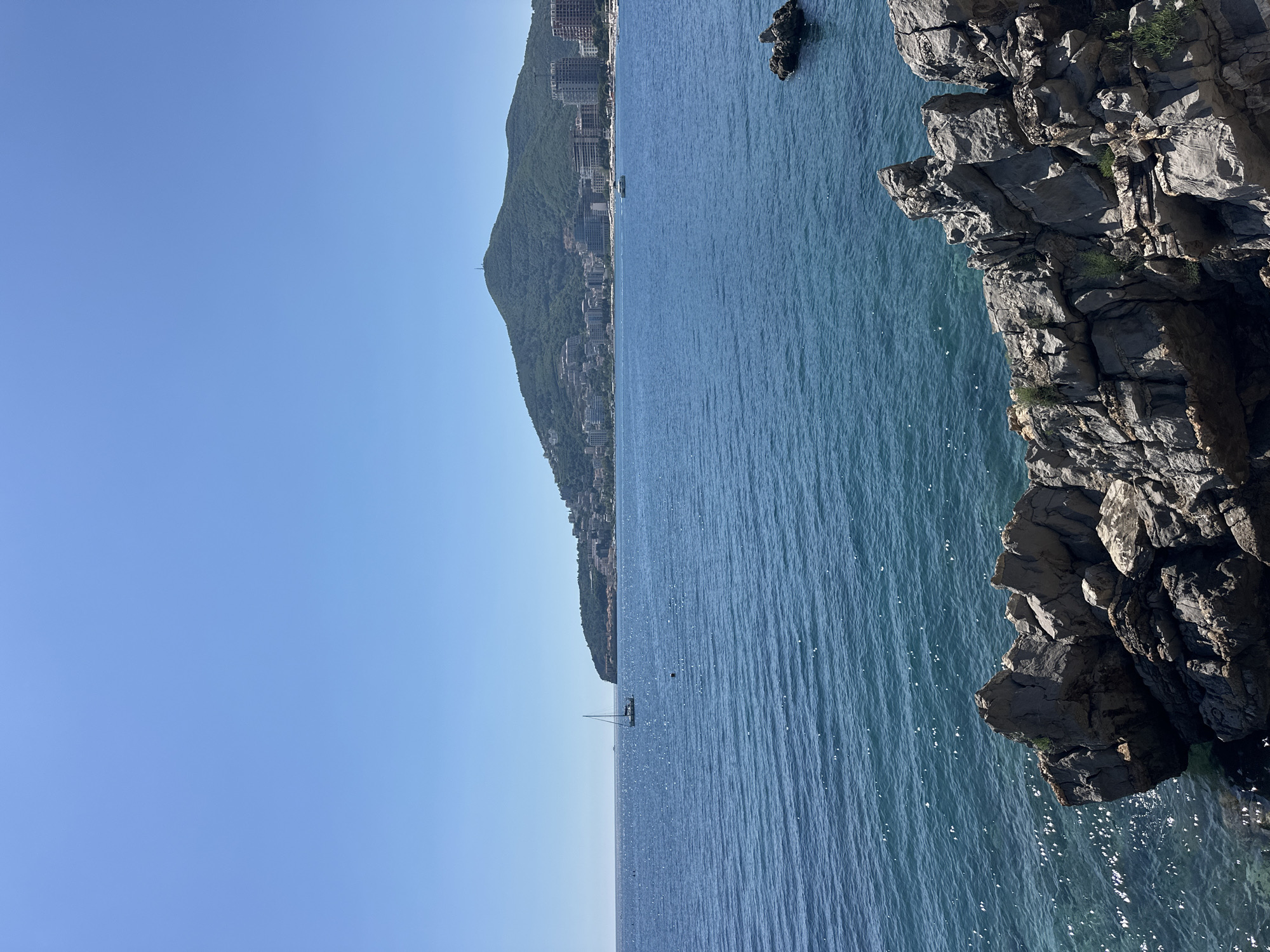 The image depicts a coastal scene in Budva, Montenegro. The view includes a stretch of coastline with a steep, rocky cliff dominating the right side of the frame. The Adriatic Sea extends towards the horizon, showing a sailboat in the distance. The mountainous terrain is lined with buildings and infrastructure. The sky is clear and blue.