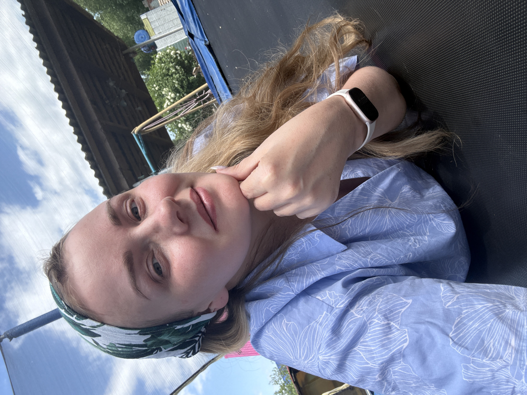 A woman is lying on a trampoline outside in Chaikovsky, Russia. She is wearing a blue floral patterned shirt and a headband. She has a fitness tracker on her wrist and is touching her face with her hand. In the background, there is a wooden structure, a blue trampoline enclosure, trees and sky.