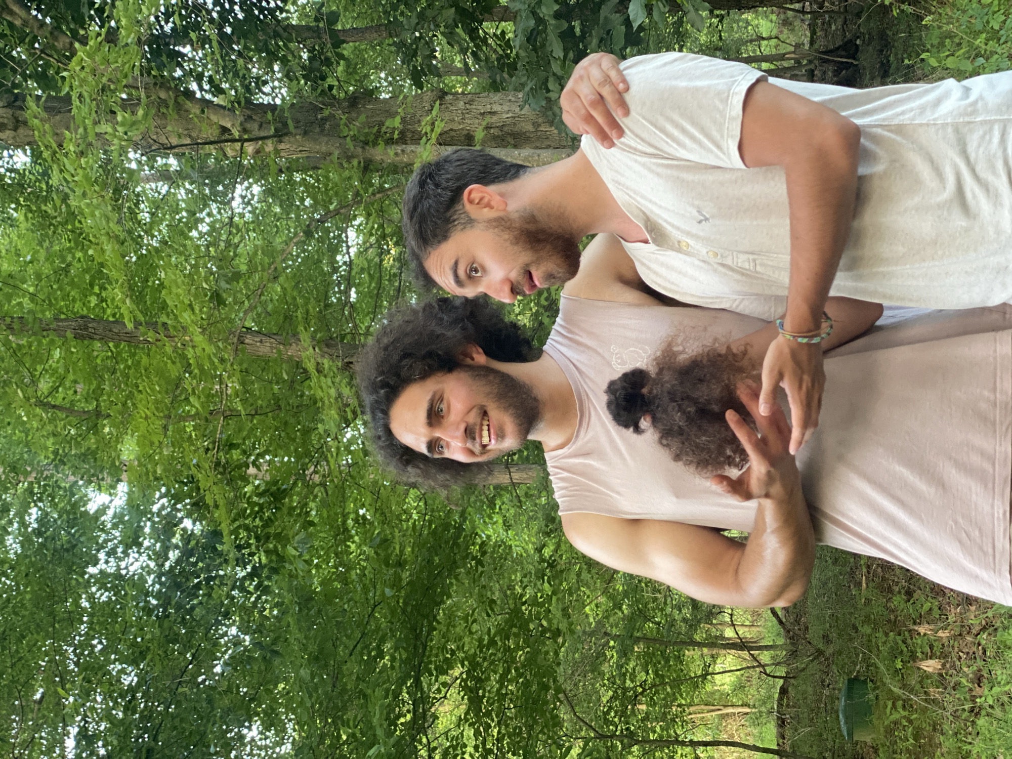 In Washington Township, United States of America, two young men are standing in a wooded area. The man on the left, with dark, curly hair and a beard, is wearing a light pink sleeveless shirt and is holding a small clump of dark hair in his hands, presenting it to the other man. The man on the right, with dark, short hair and a beard, is wearing a light-colored henley shirt and has his arm around the other man's shoulder. They are both looking towards the left of the frame, with expressions of interest and amusement. The background is filled with lush green trees and foliage, suggesting an outdoor, natural setting.