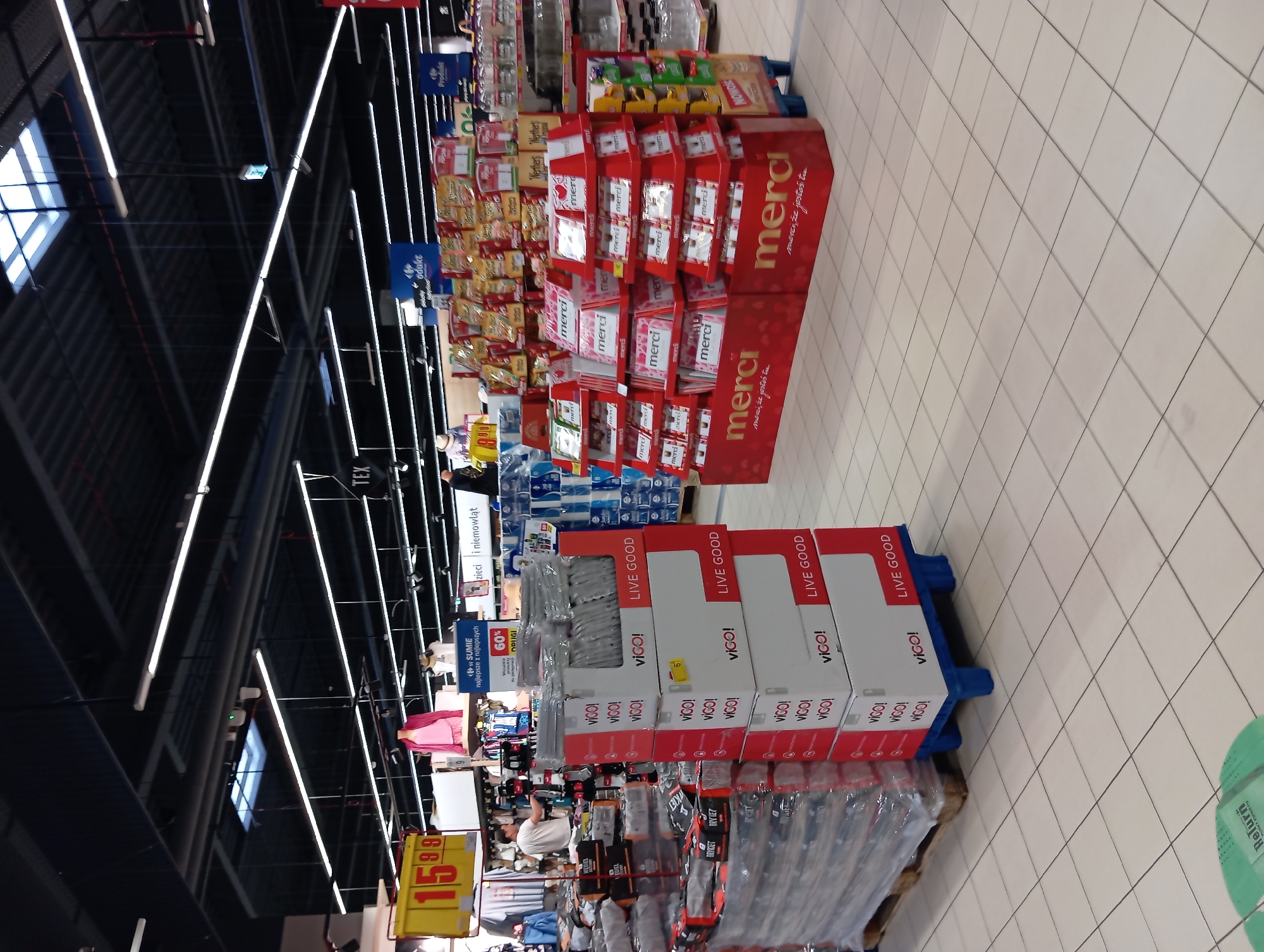 The image depicts the interior of a brightly lit supermarket, likely a Carrefour based on signage. The flooring is tiled in a light beige color, and the ceiling features a dark metal structure with numerous fluorescent lights and ventilation systems. In the foreground, a stack of boxes with red and white branding, featuring the word "VIGO!" and "LIVE GOOD" prominently, are arranged on a blue plastic pallet. To the left, several large pallets wrapped in plastic are visible, containing dark-colored products. Further back, a display of red tiered stands holds numerous smaller boxes branded with "merci". A variety of other products, including bags of snacks and bottles, are visible on shelves in the background. There are no visible people interacting with the merchandise. The overall scene suggests a typical retail environment within a commercial establishment in Wołomin, Poland.