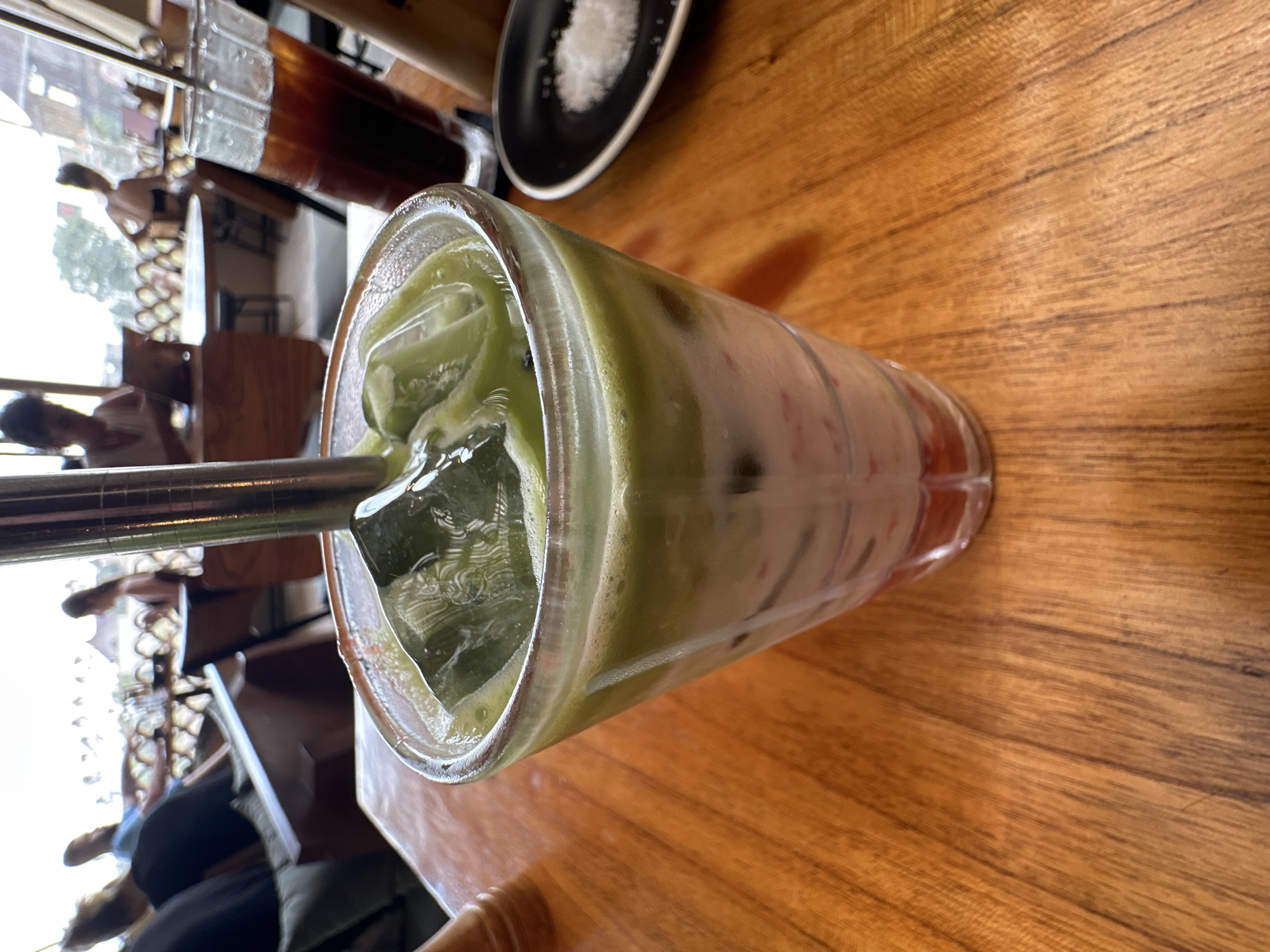 A close-up, high-angle shot depicts a tall glass of iced matcha latte on a wooden table, likely at a café. A metal straw protrudes from the top of the glass. In the background, partially visible, are other tables, chairs, and people. A small black plate with white powder, possibly sugar or salt, is also visible in the background. The image suggests an urban setting, potentially in Indonesia, although the precise location is unknown.