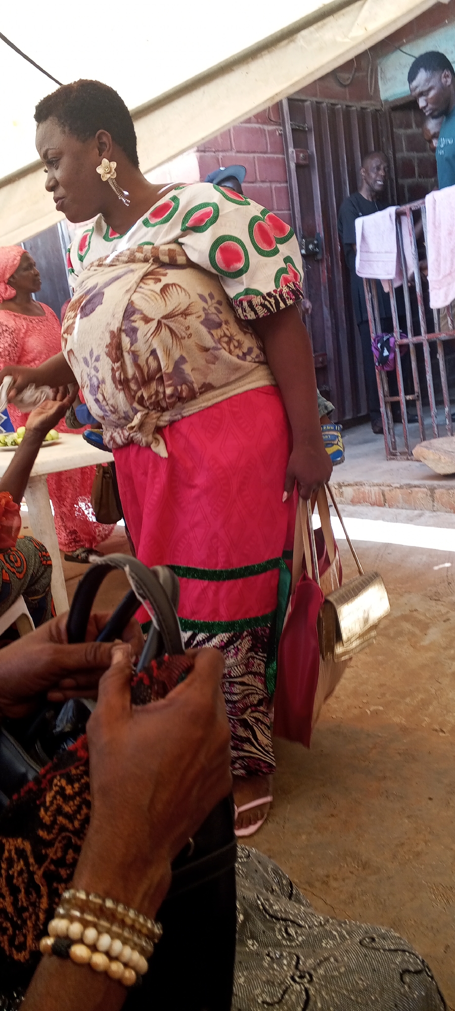 The image shows a woman in Unknown, Unknown, wearing a colorful outfit including a white top with red and green circular designs, a floral-patterned wrap, a pink skirt with green trim, and patterned leggings. She is carrying a golden clutch purse and a red bag. In the background, there are people standing near a building with barred windows and laundry hanging to dry. Another woman, wearing a pink headscarf and dress, is partially visible to the left. A person in the foreground is holding a black bag and wearing multiple bracelets.