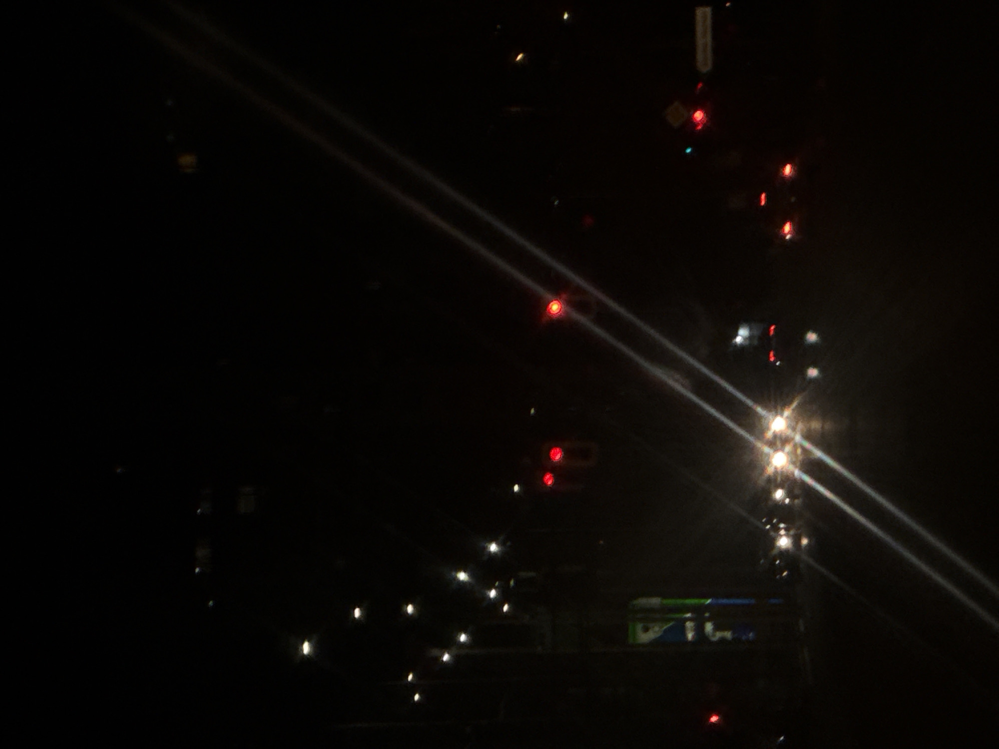 The image captures an urban street scene at night, likely from within a vehicle, characterized by darkness punctuated by artificial light sources. Multiple bright streetlights are visible on the left side of the frame, accompanying a tall, vertical commercial sign predominantly blue and green. In the immediate foreground-center, a line of approximately 5-7 vehicles displays active, circular white headlights, suggesting they are facing the viewer and potentially stopped. Two distinct red traffic signal lights are present in the upper-left and upper-center sections of the frame. To the right, several vehicles are visible from the rear, exhibiting red tail lights. Prominent diagonal lens flares, appearing as two parallel white streaks, traverse the image from bottom-left to top-right, originating from the headlights. No individuals are visible. The exact location within the city of Unknown, Unknown, is not discernible.