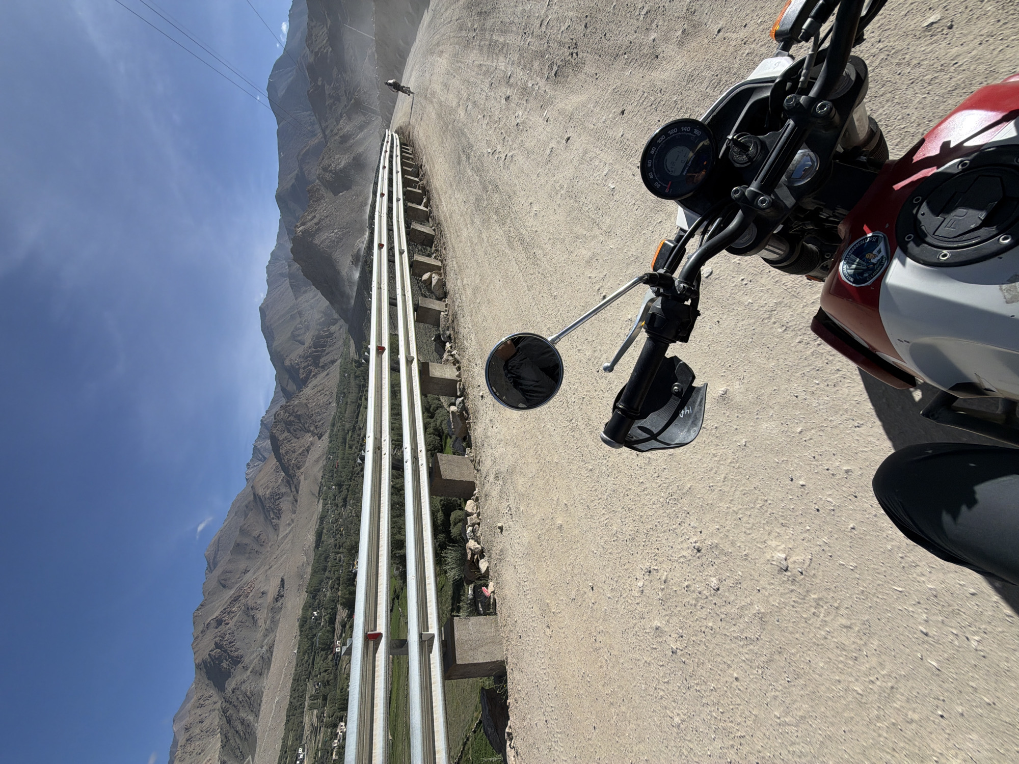 An over-the-shoulder view captures a motorcycle rider's perspective on a dusty, unpaved road winding through a mountainous landscape. The motorcycle's handlebars, dashboard, and a portion of its red and white fuel tank are visible in the foreground. The road continues ahead, curving to the right, with a white metal guardrail bordering its left side. Beyond the guardrail, a lush green valley with scattered buildings and trees stretches out, leading to the imposing, barren mountains that dominate the background under a clear blue sky. In the distance, another motorcyclist is seen on the road, kicking up dust. The scene suggests a journey through the rugged terrain surrounding Leh, India.