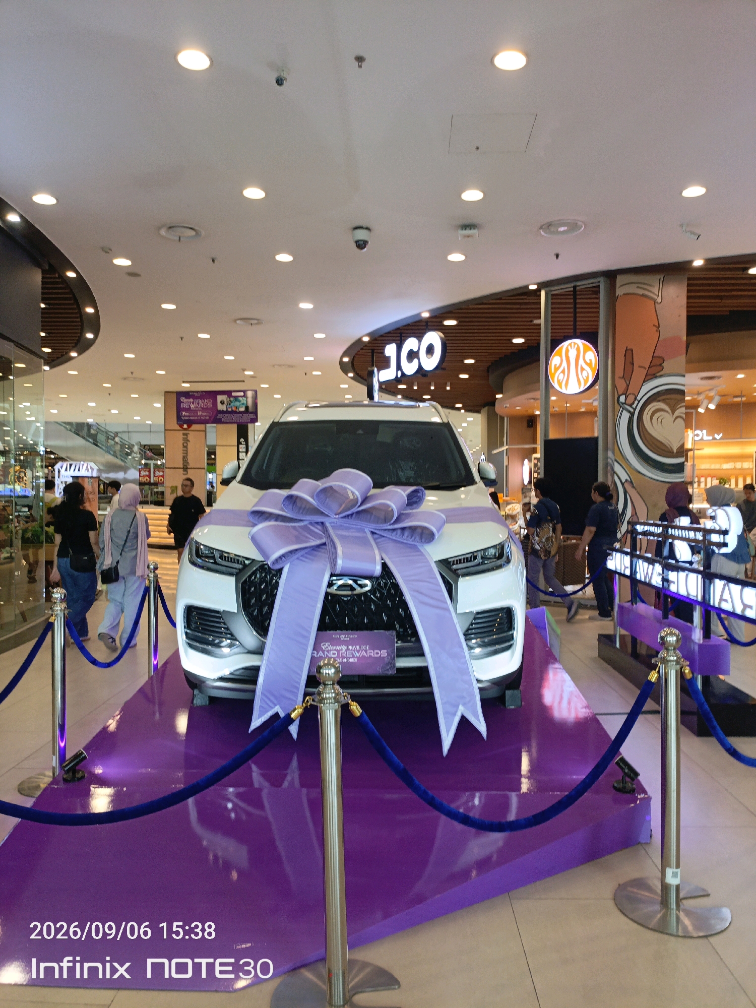 A white SUV, adorned with a large purple bow, is displayed on a raised purple platform in what appears to be a shopping mall. The car is positioned centrally, with its front grille and headlights prominently visible. The mall environment is characterized by a high ceiling with numerous circular light fixtures, some of which are illuminated. In the background, several individuals are walking, and there are signs for shops, including "J.CO" and another with a stylized logo resembling a coffee cup. A banner above the car reads "Eternity Privilege of Grand Rewards." Blue velvet ropes on silver stanchions cordon off the display area. The date stamp on the image indicates "2026/09/06 15:38" and the device used is an "Infinix NOTE 30". This scene is likely located within the AEON Mall Sentul City in Babakan Madang, Indonesia, given the branding and general appearance of a modern Indonesian shopping mall.