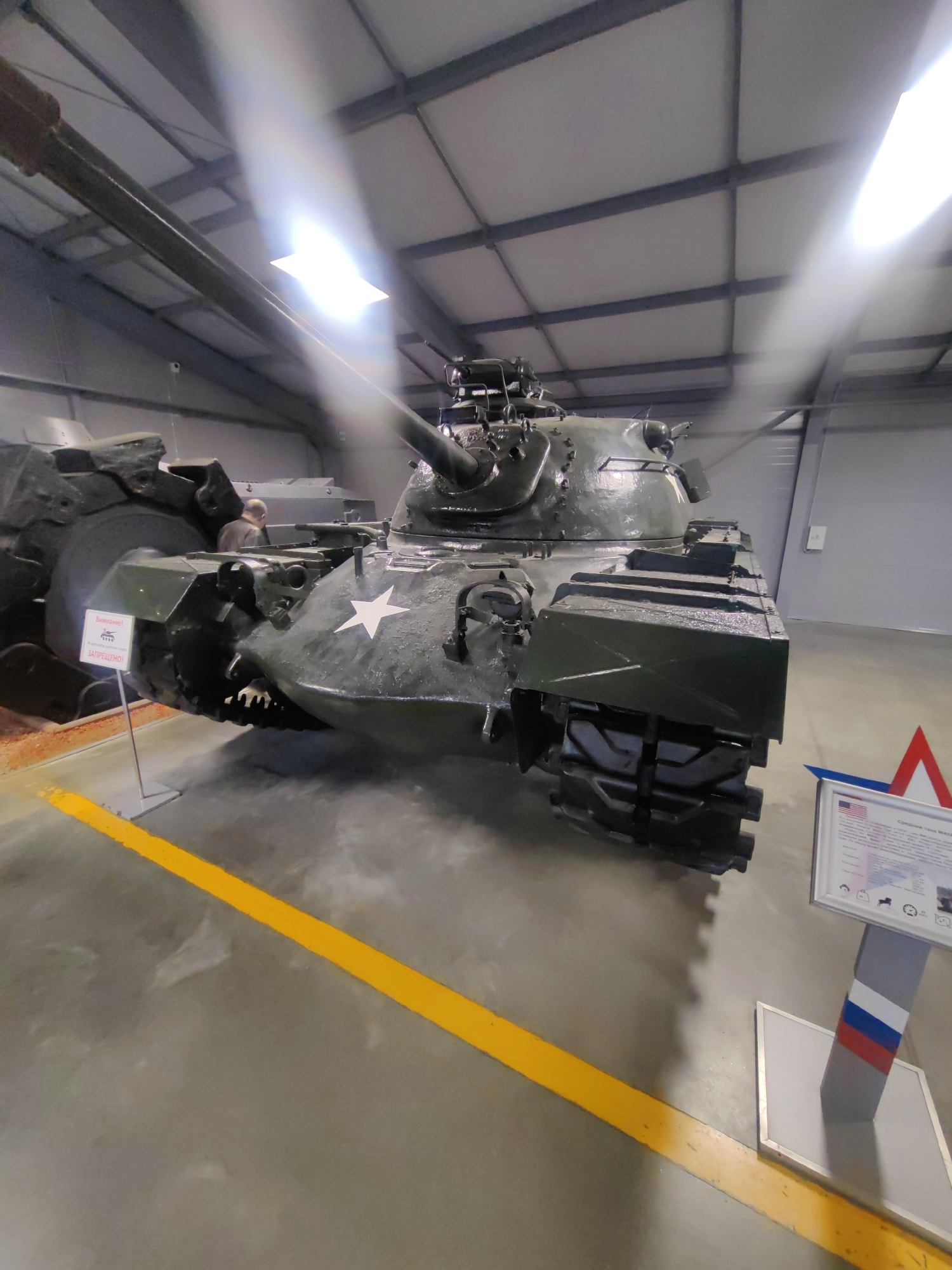 The image shows a M4A2 Sherman medium tank displayed indoors. The tank is painted olive drab and features a white star emblem on its turret. The setting appears to be a museum or exhibition hall, with a high ceiling, metal beams, and bright overhead lighting that causes lens flare. A person is visible in the background, looking at the tank.
To the left of the tank, there is a sign with red text that reads "Внимание! РАБОТАЕТ ЗАПРЕЩЕНО!" which translates to "Attention! Operation is prohibited!". This sign indicates that the tank is a static display and not operational.
In the foreground, a bright yellow line runs across the floor, marking a boundary around the exhibit. To the right of the tank, a display stand features a framed informational plaque with text and icons, along with the flags of the United States and Russia, suggesting the tank has historical significance in both countries. The overall impression is of a well-maintained historical exhibit.