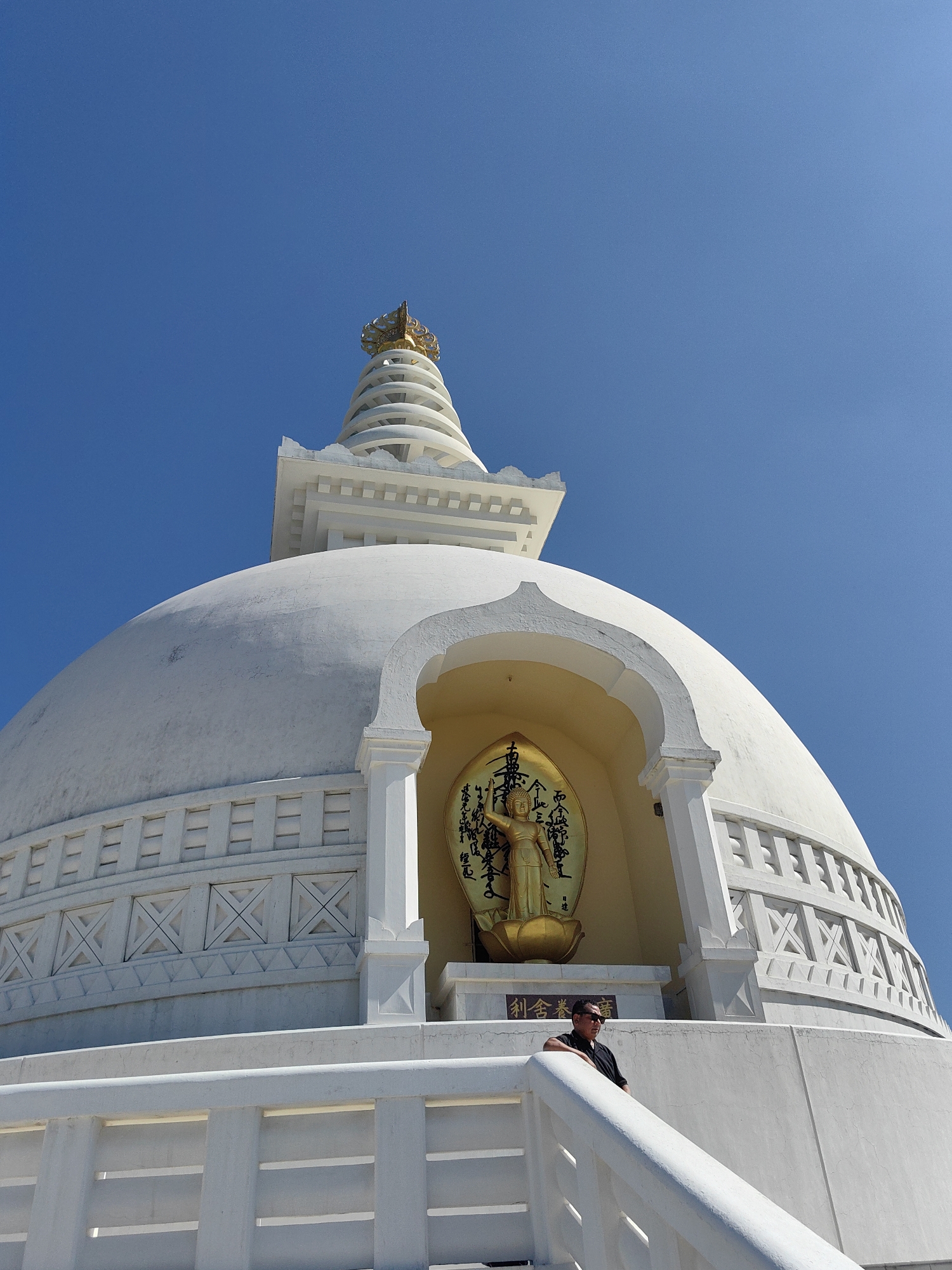 An extreme low-angle, medium shot captures the upper portion of a white stupa against a clear, bright blue sky. The stupa's dome is prominent, with a decorative, latticed band encircling its base, featuring alternating square and triangular patterns. Above the dome, the stupa tapers into a series of stacked circular platforms, culminating in a golden, multi-tiered finial. Centered on the dome is a golden, arched alcove containing a golden statue of Buddha standing on a lotus flower. Behind the Buddha is a circular, golden disc inscribed with what appear to be calligraphic characters. A man wearing sunglasses and a black shirt stands on a white, tiered platform to the right of the alcove, leaning on a white railing. The scene is set outdoors, under a sunny sky, with no other structures or natural elements visible. The location is identified as Lumbini Bazaar, Nepal.