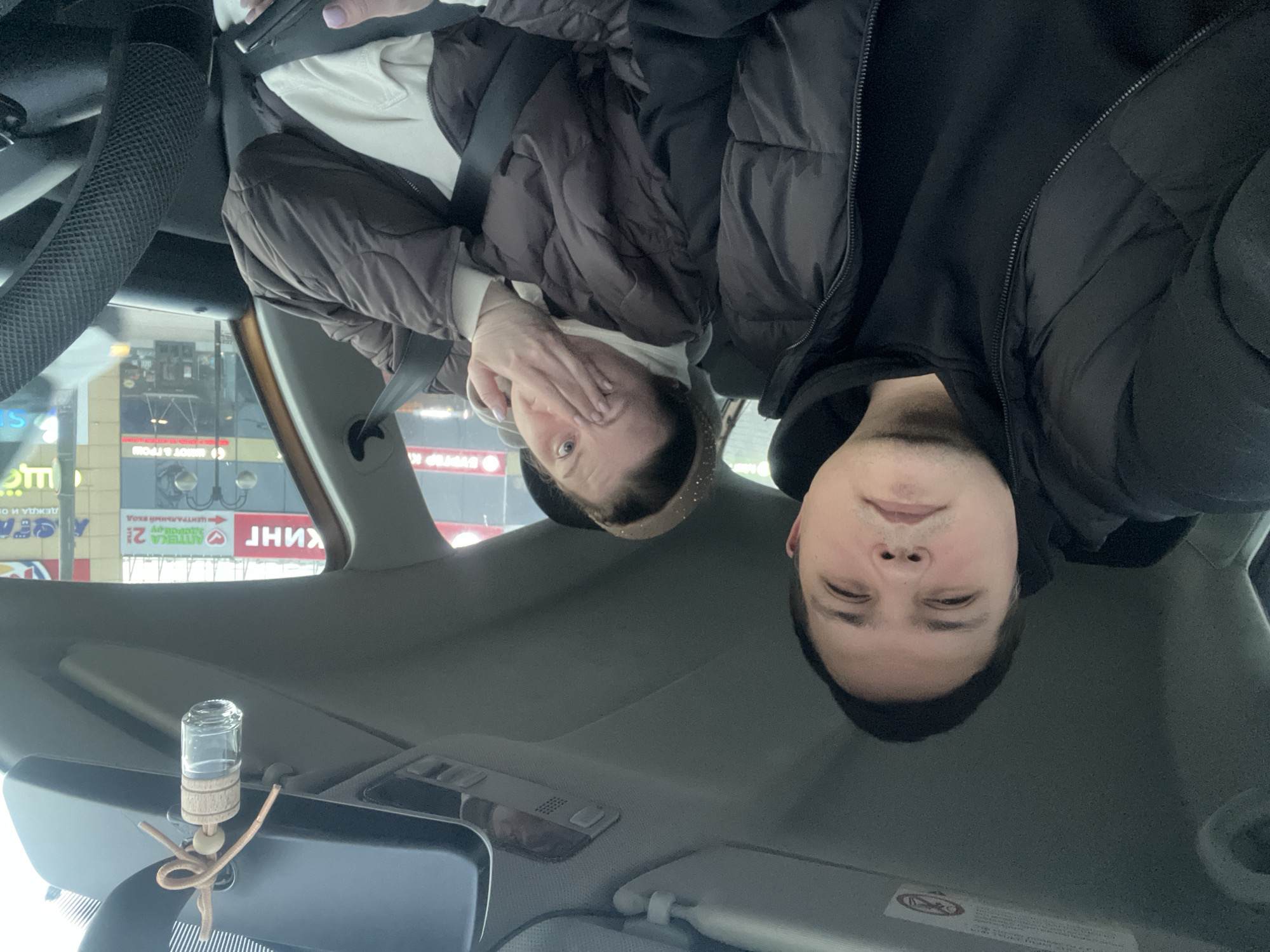 The image captures two individuals in a car, likely in the city of Klin, Russia. A woman in a brown puffer jacket with a white top partially covers her face with her hand. A man in a black puffer jacket is next to her. Both are wearing seatbelts. A car freshener hangs from the rearview mirror. Outside the car window, signs written in Russian are visible, including "Бургер Кинг" and "Аптека 21" (Pharmacy 21).