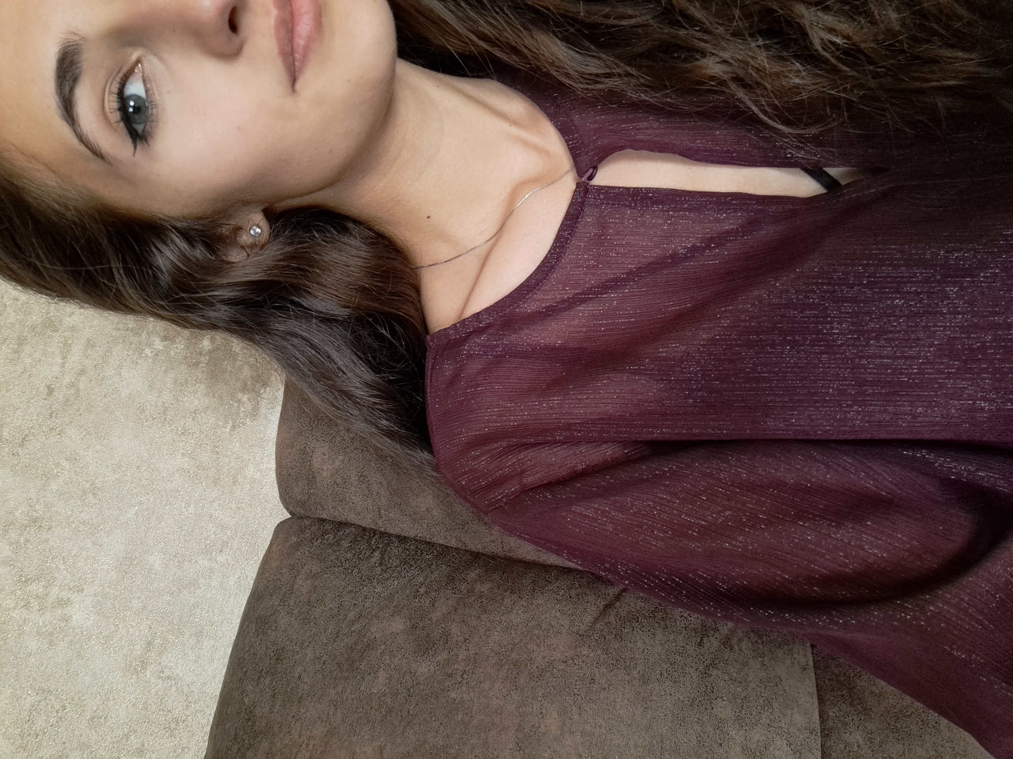 A young woman with long, wavy brown hair, light skin, and green eyes is lying on a brown suede-like couch. She is wearing a sheer, maroon-colored blouse with subtle metallic threading. A silver necklace is visible around her neck. She has a stud earring in her left ear. The lighting appears soft and diffused. It's not possible to determine the location in Negovan, Bulgaria from the image.
