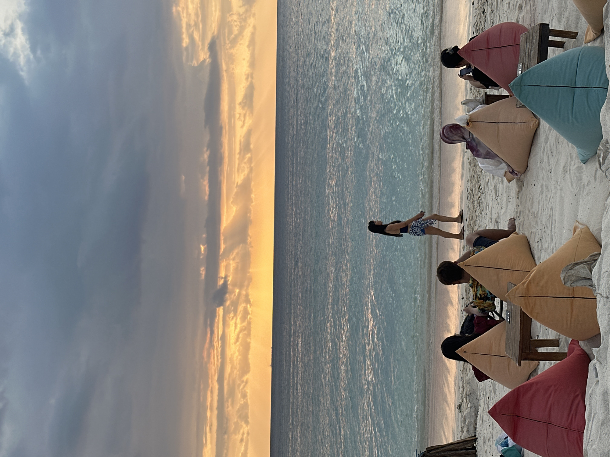 The image captures a beach scene during sunset. The sky is divided, with a dark, cloudy area on the left and a bright, orange-hued sunset on the right. The ocean stretches across the middle, reflecting the colors of the sky.
Several individuals are present on the beach. Some are lying down, while others are sitting on colorful triangle-shaped cushions. The cushions are arranged around small wooden tables. People appear to be looking at the water.
The location appears to be a beach in an unidentified city in Indonesia.