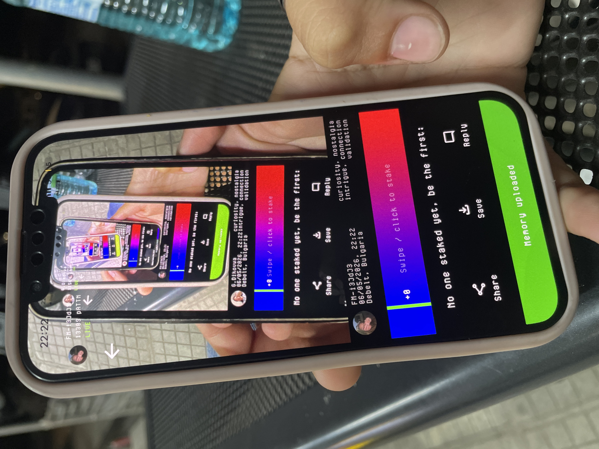 A person's hand holds a light-colored smartphone, displaying an application interface, against a blurred background. The phone, encased in a beige or light pink shell, shows the time "22:22" in the top left corner of its screen. The application features a recursive image, where a smartphone displaying the same application is shown within the current screen, creating a visual loop.
On the outermost displayed screen, a small circular profile picture is visible in the header, with text below identifying the content creator as "FM-i3JDj3" and stating the timestamp "06/05/2026, 22:22" and location "Debelt, Bulgaria," followed by "13389 PNTTM LIVE." White arrows point inwards from the left and right edges. Below this header is the recursively nested image. Below the main content, a horizontal bar features blue and red sections with "+0 Swipe / click to stake" text. Further down, "No one staked yet, be the first:" is displayed, followed by icons for "Share," "Save," and "Reply." A prominent green banner at the bottom reads "Memory uploaded."
Within the first nested screen, details for "G.Dikouva" are visible, along with the same timestamp and location "Debelt, Bulgaria." Associated keywords "curiosity, nostalgia intrigue, connection validation" are listed. This screen also shows the blue and red staking bar and the "No one staked yet" prompt. The innermost nested screen displays a colorful, abstract pattern.
The hand holding the phone has light skin and visible, neatly trimmed fingernails with a faint pink or clear polish. In the blurred background, a translucent blue plastic water bottle with a white cap, partially filled with liquid, is visible on the right. The phone rests partially on a dark, perforated or gridded surface.